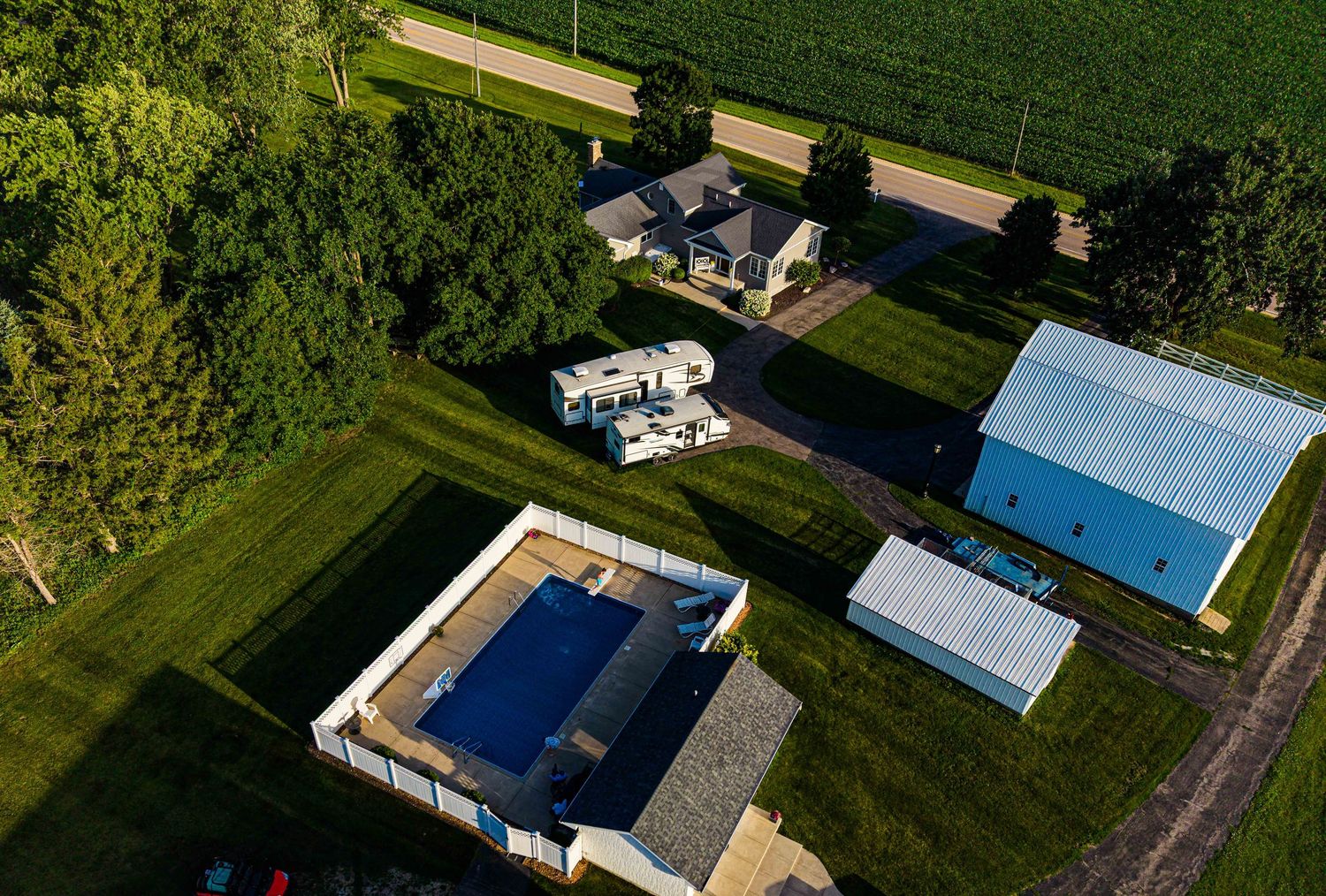 Aerial view of a rural property featuring a farmhouse, blue metal barn, pool, and lush green fields.