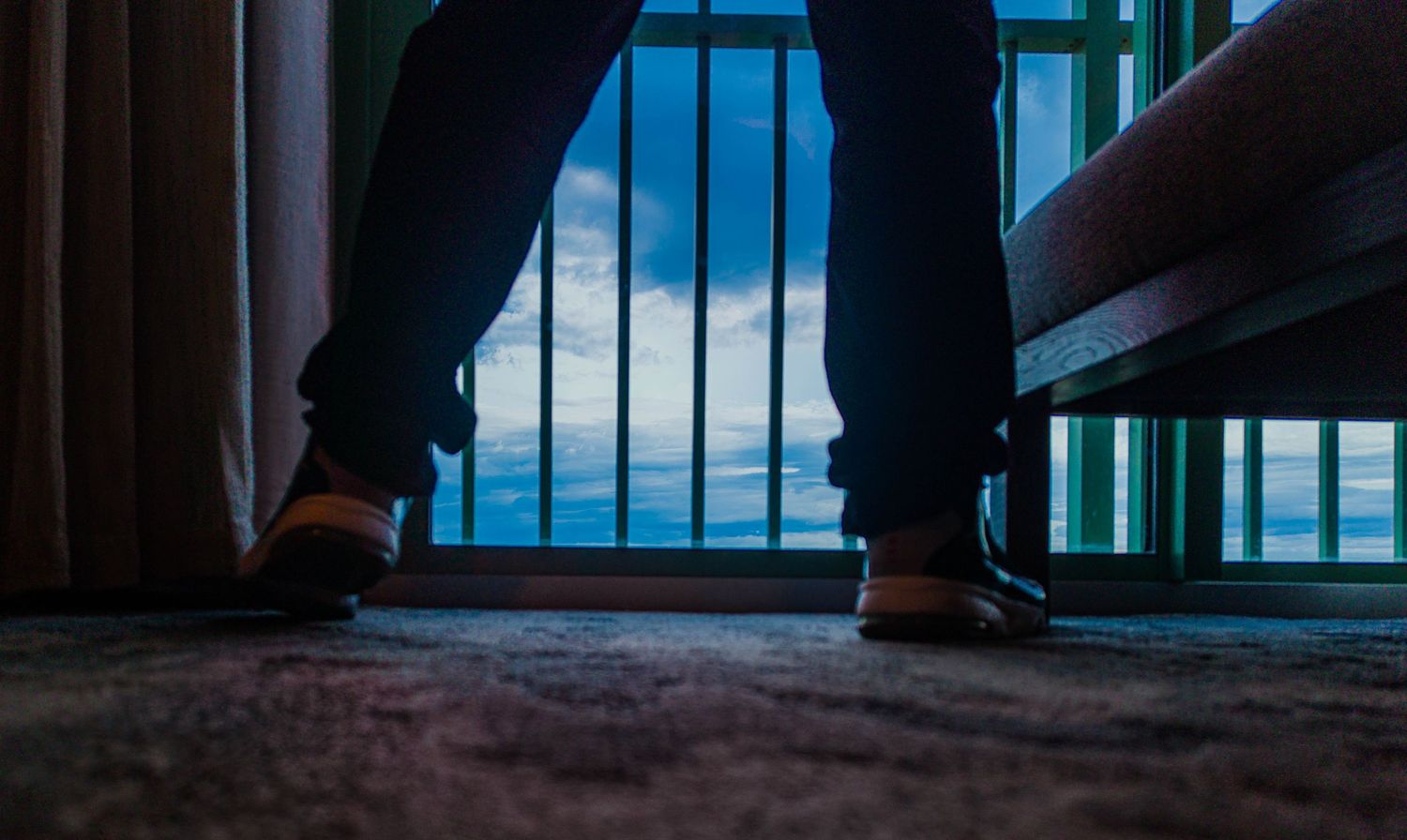 Person standing in dark room facing barred window with blue sky view, wearing brown shoes and dark pants.