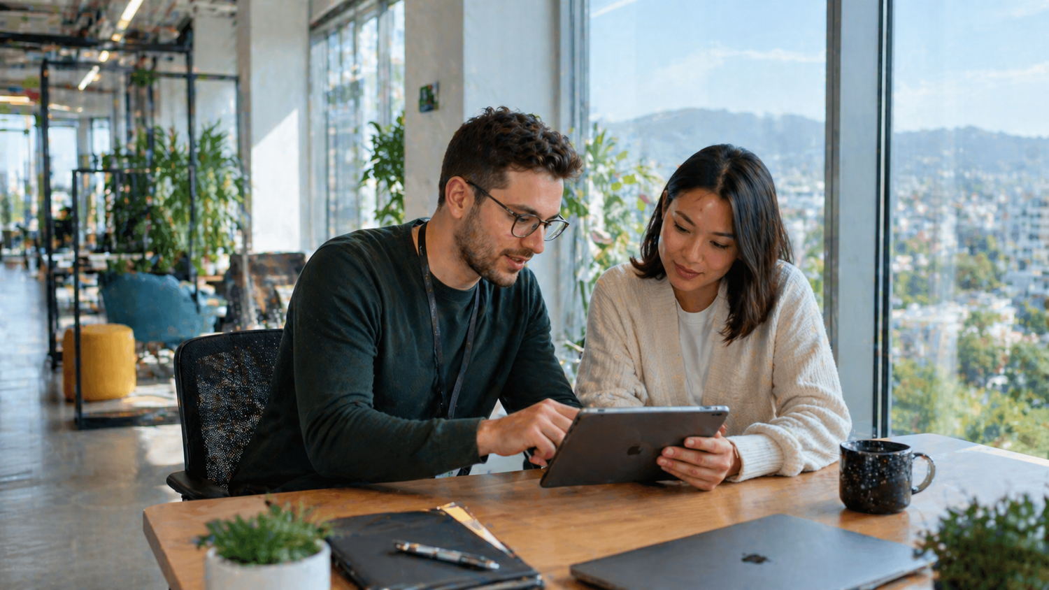 Two professionals collaborating on a tablet in a modern office with large windows and a city view.