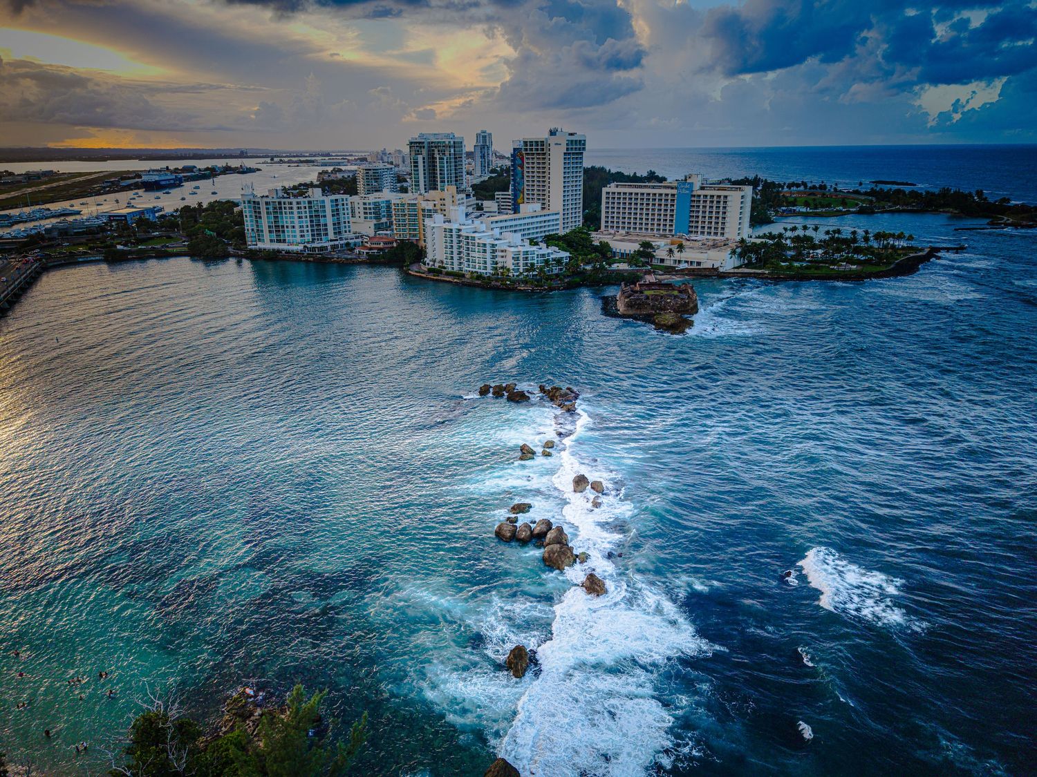 Aerial panorama of coastal cityscape with high-rise buildings and crashing waves against rocky shoreline at sunset.