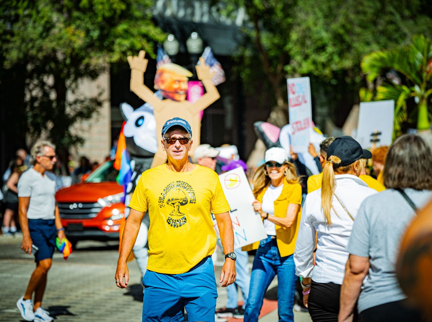 Colorful crowd of protesters marching along a tree-lined street during a daytime demonstration.