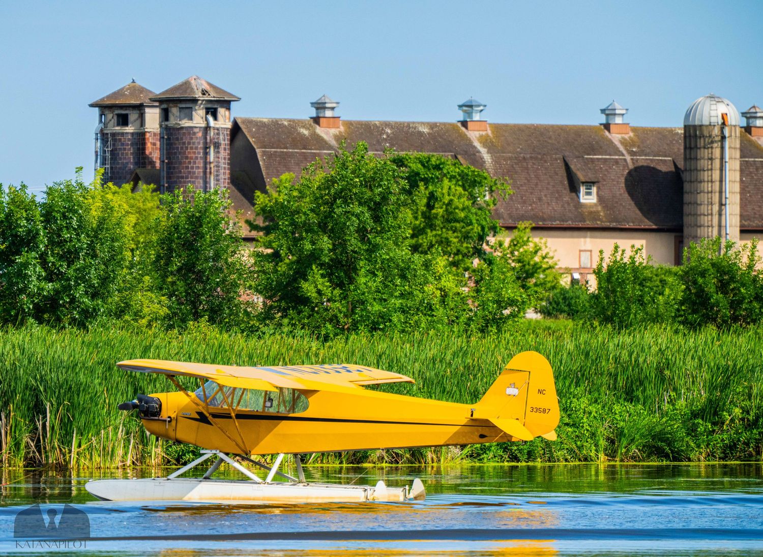 Yellow seaplane floats on lake water against backdrop of large barn with silos.