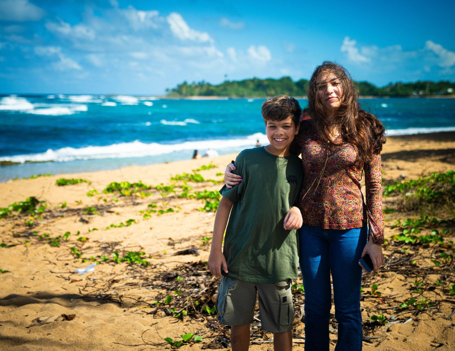 A couple stands together on a scenic beach with blue ocean waters and tropical coastline in the background.