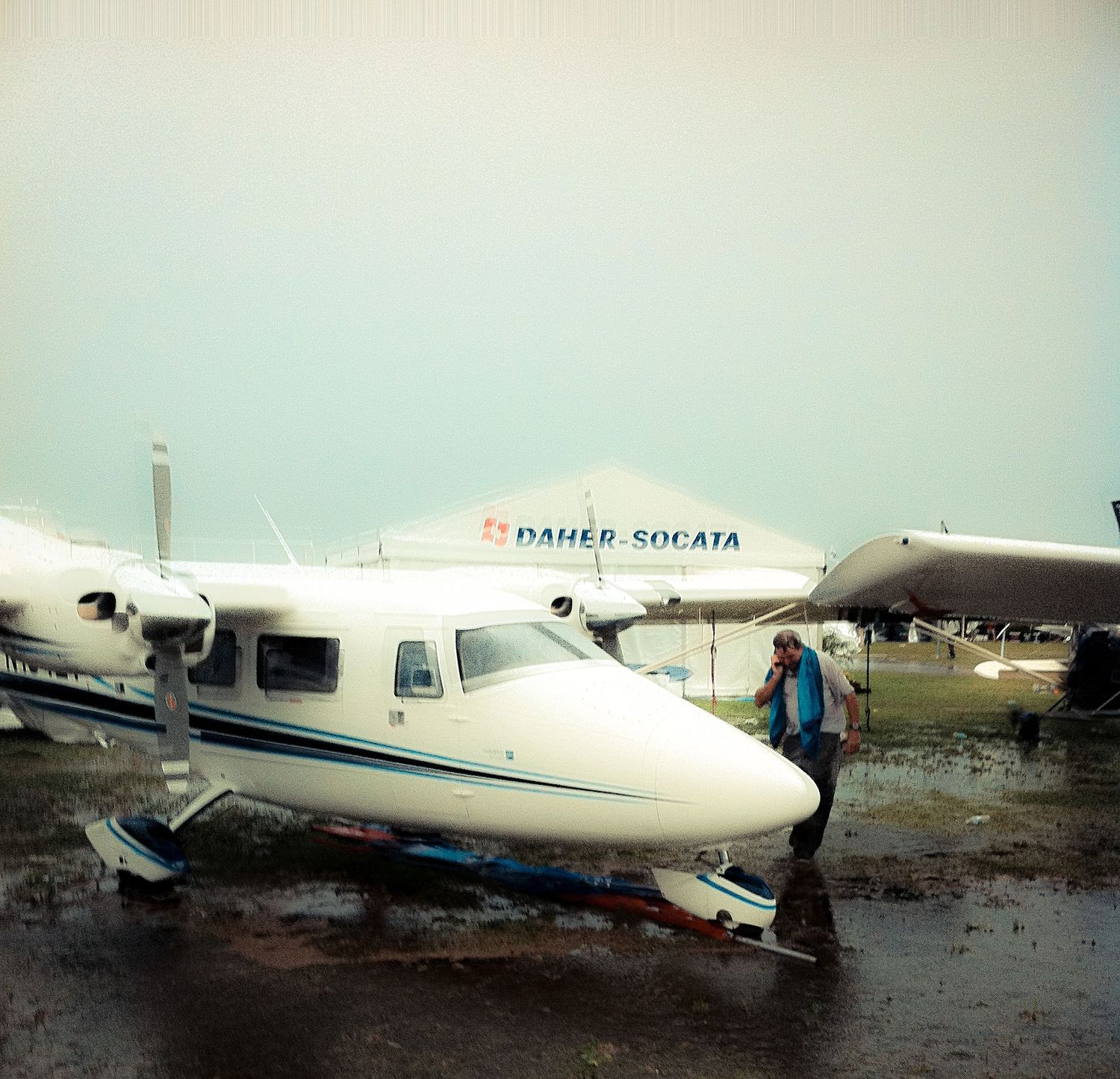 A small white passenger aircraft parked on wet tarmac with a larger commercial plane visible in the background.
