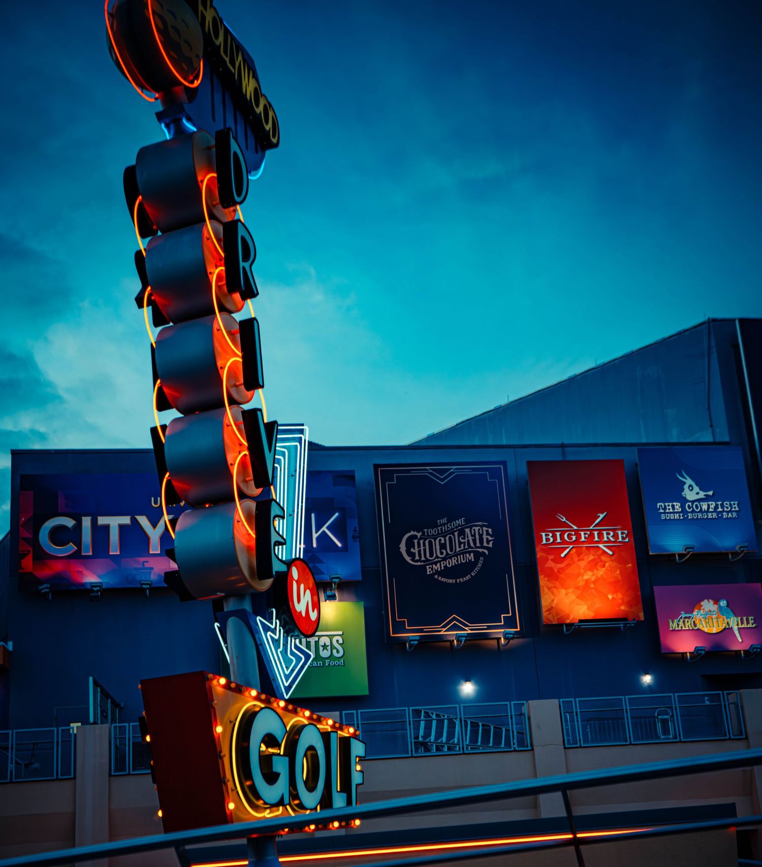 Illuminated vintage neon golf sign glows against twilight sky with City Walk and entertainment venue signage in background.