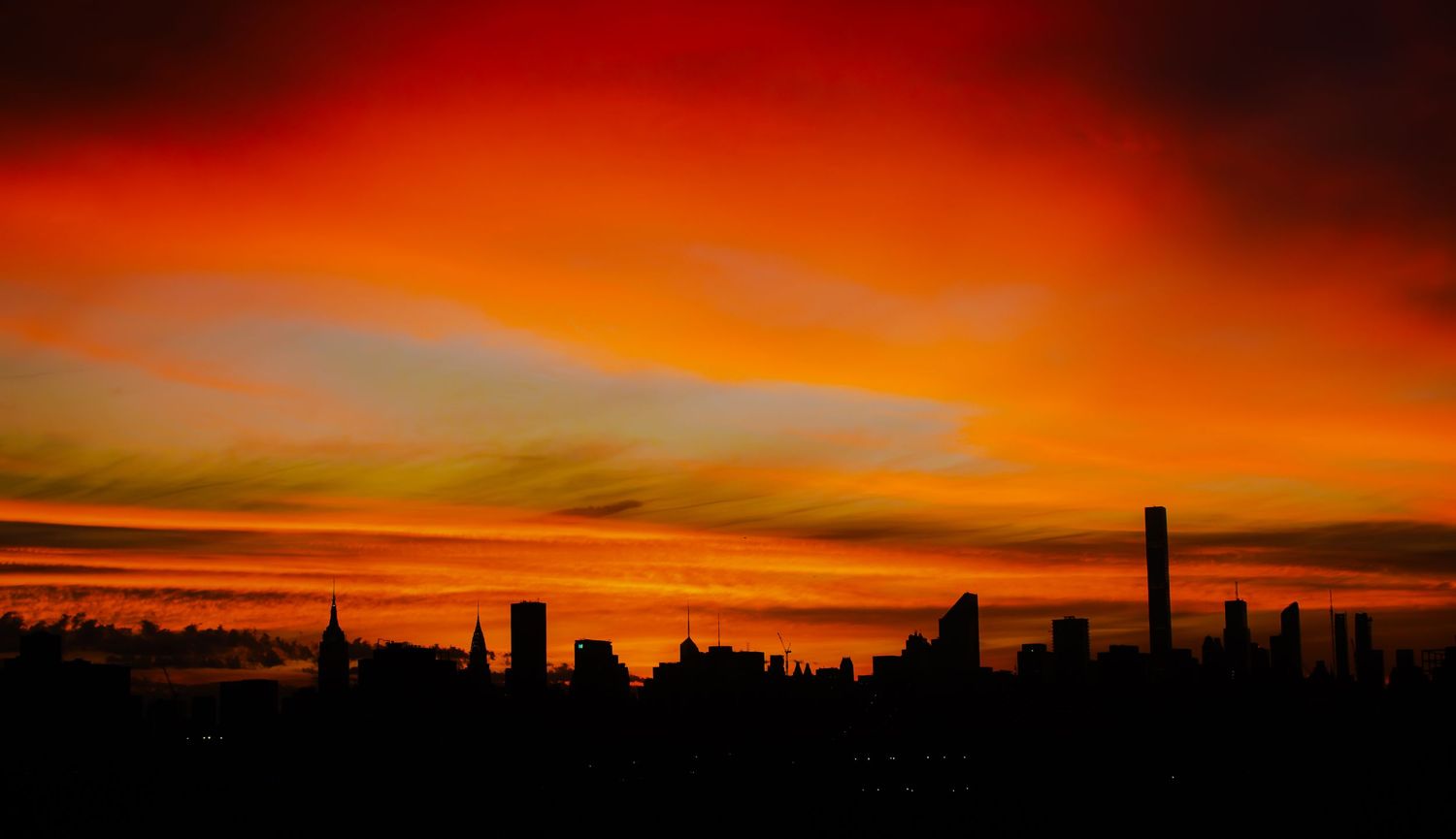 Dramatic orange and red sunset sky with silhouetted buildings and trees creating a striking panoramic view.