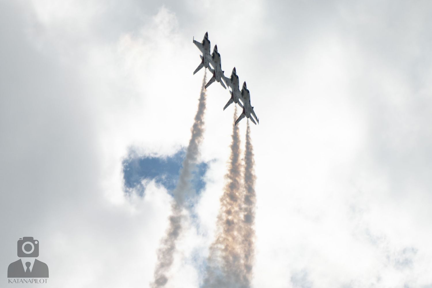 Four military jets fly in tight diamond formation, trailing white smoke against a cloudy sky at an airshow.