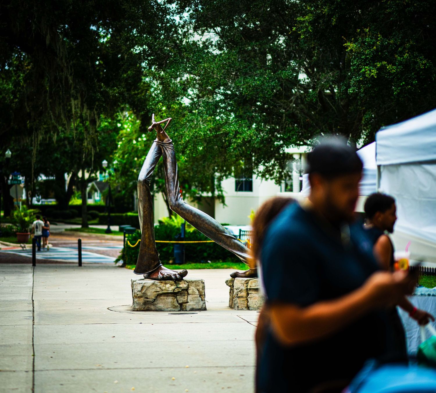 Outdoor plaza with a tall bronze cowboy statue on stone base, surrounded by trees, with blurred people walking nearby.