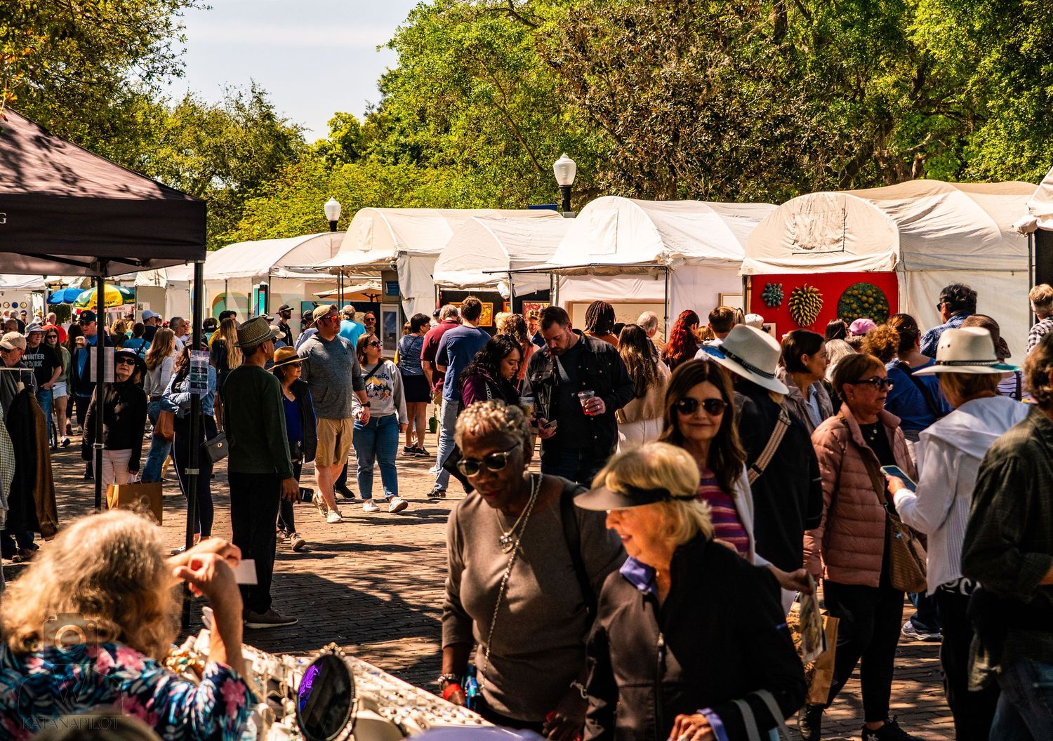 Busy outdoor market with white vendor tents and crowds browsing merchandise.