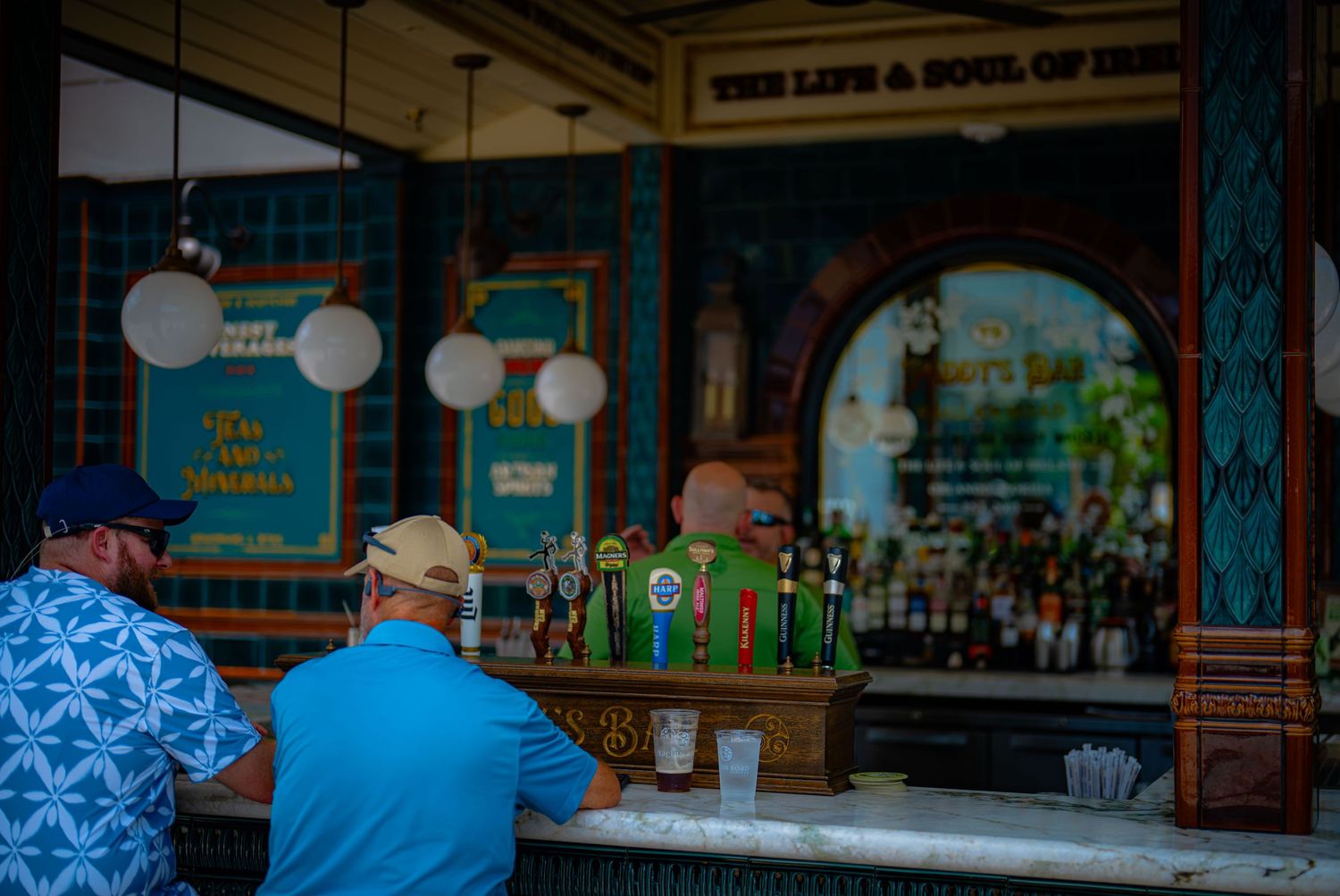 Two men sit at a colorful outdoor bar with draft beer taps, beneath a sign reading The Life and Soul of Ireland.