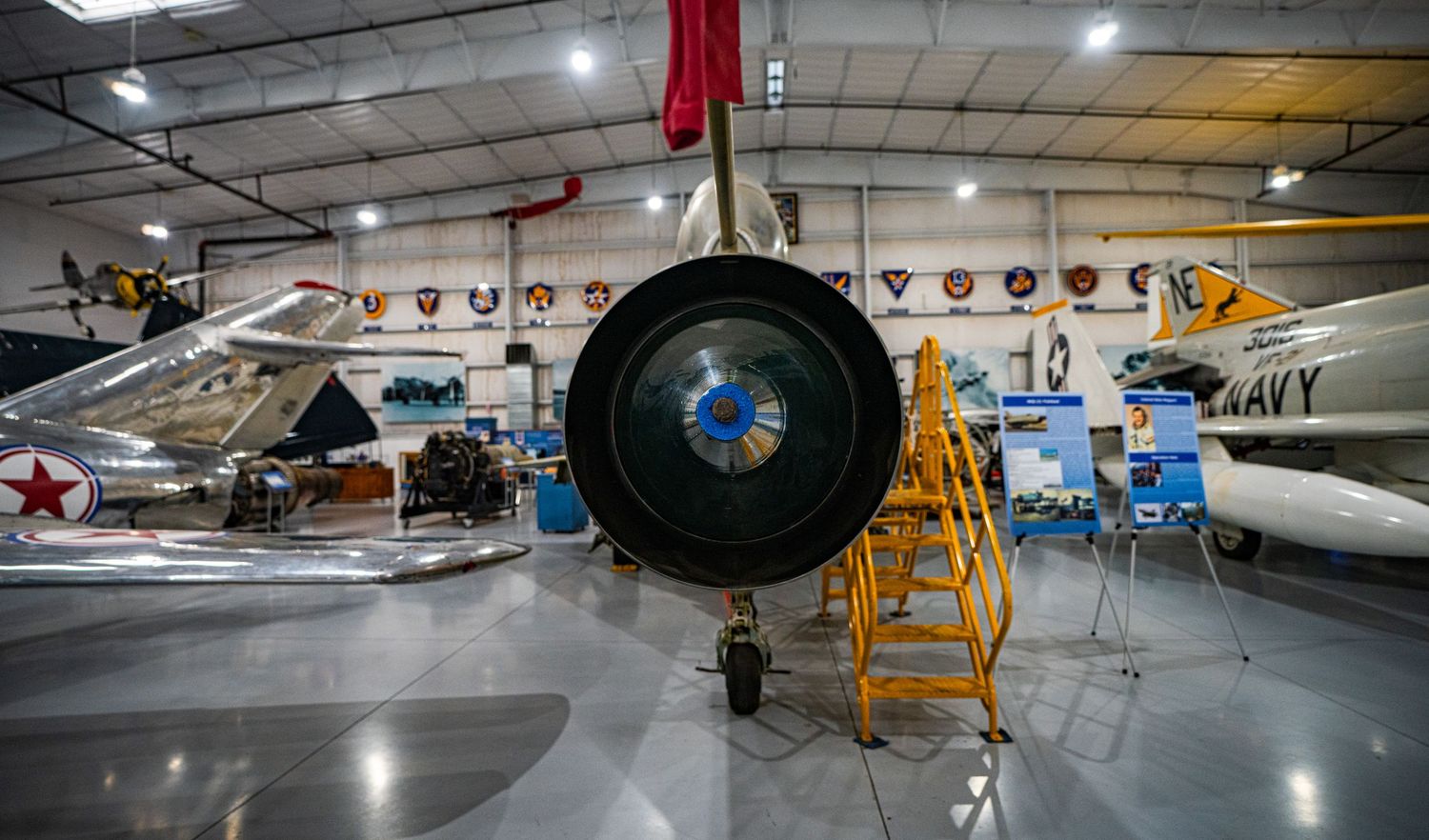 Military aircraft displayed in a museum hangar, featuring a jet engine close-up with Navy planes visible in the background.