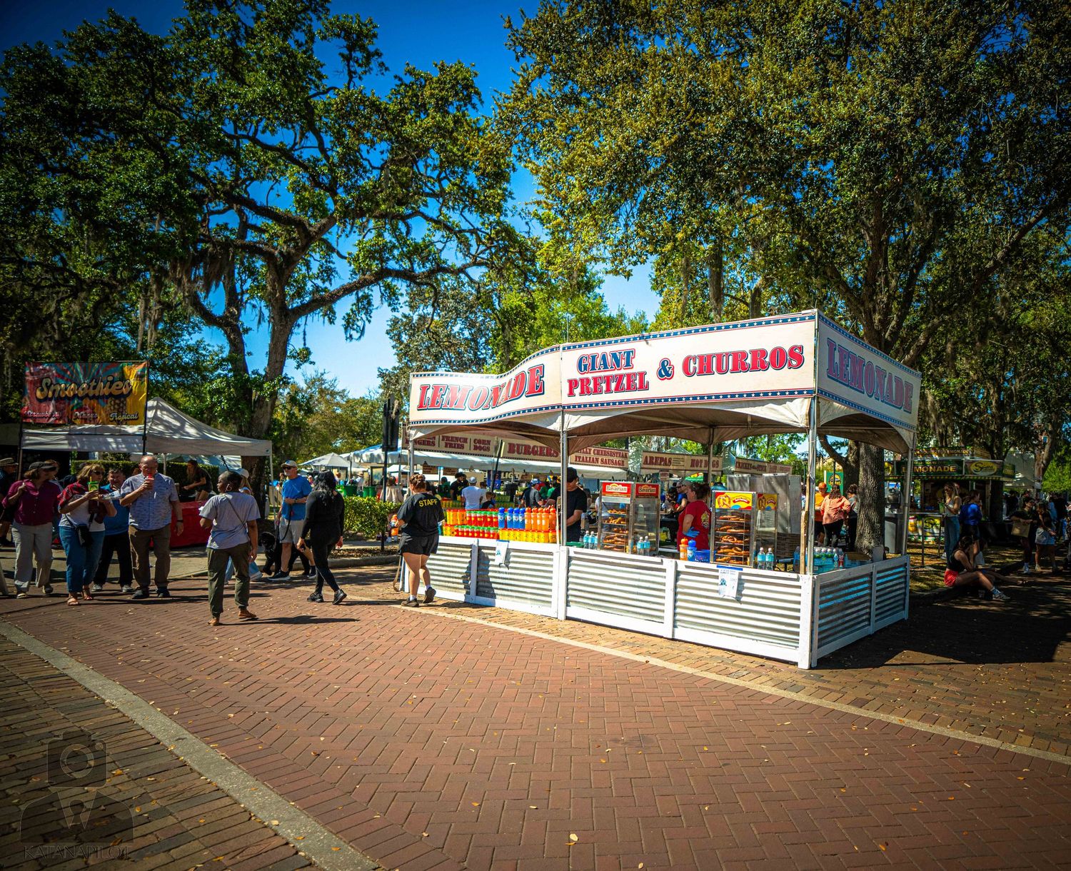 Panoramic view of a sunny outdoor fairground with food vendor stalls and visitors walking along a path.