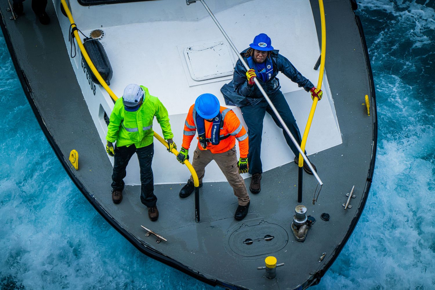 Three crew members in colorful safety gear work on the bow of a vessel surrounded by rough ocean waves.
