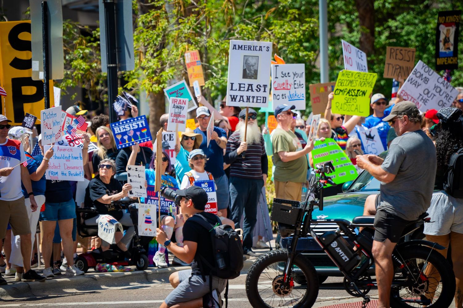 Street protest scene with demonstrators holding various signs while some participants sit on motorcycles.