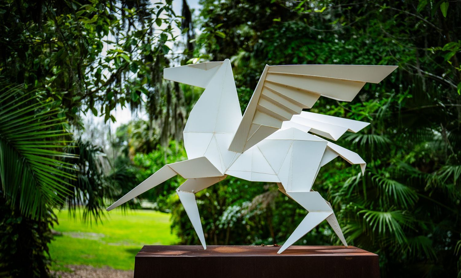 Modern white pegasus sculpture with angular design set against green foliage.