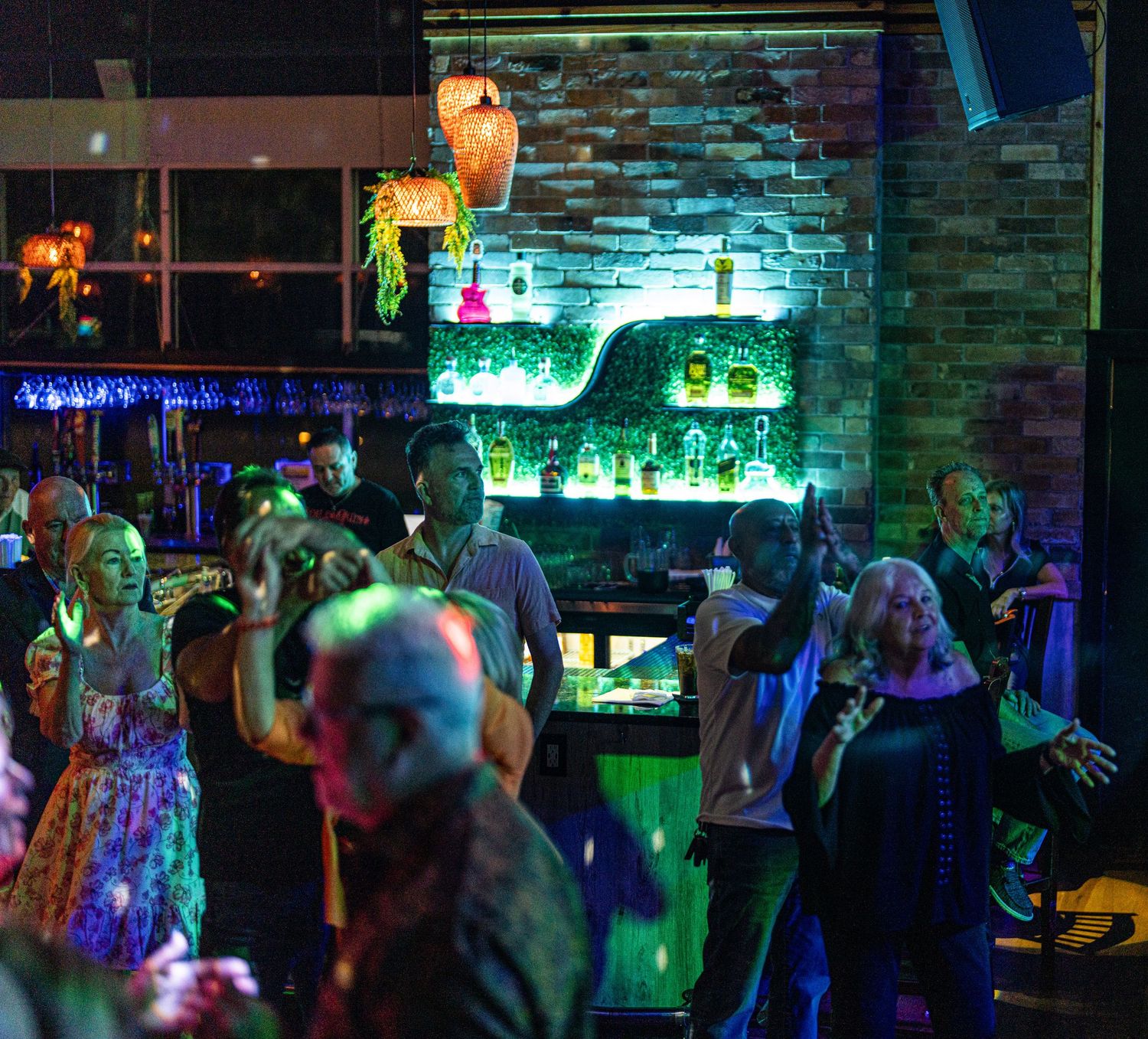 Lively nightclub bar scene with neon green lights illuminating liquor shelves while guests dance and socialize in vibrant atmosphere.