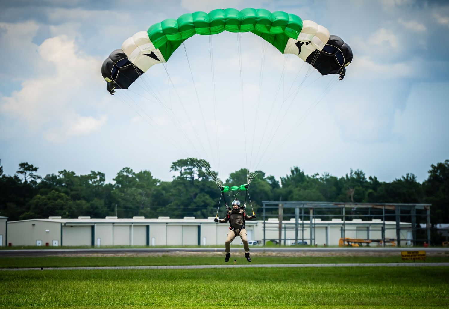 Green parachute canopy spread wide during final landing approach over an airfield.