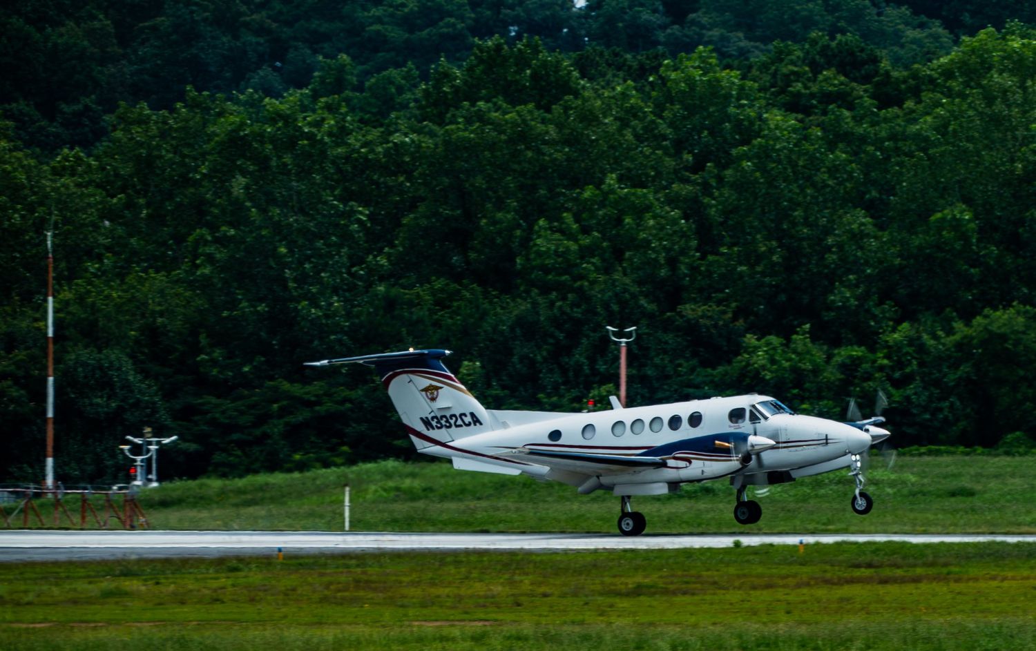 Small twin-engine propeller aircraft taxiing on runway surrounded by lush green trees at a regional airport.