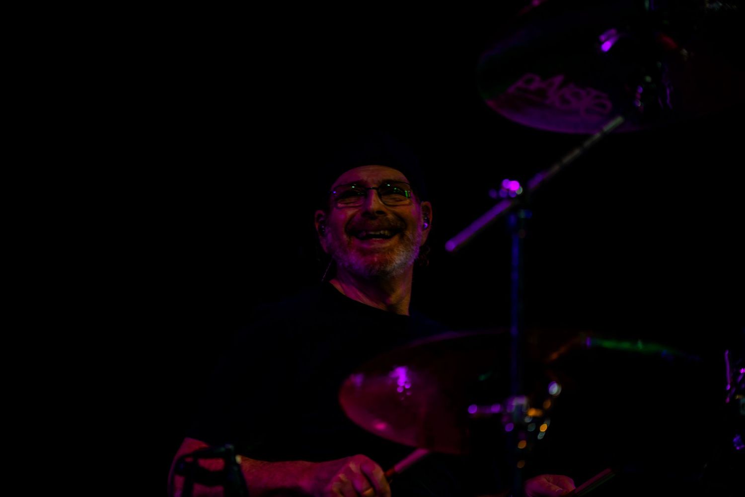 Smiling drummer performing on stage under purple stage lighting with cymbals visible in dark concert setting.