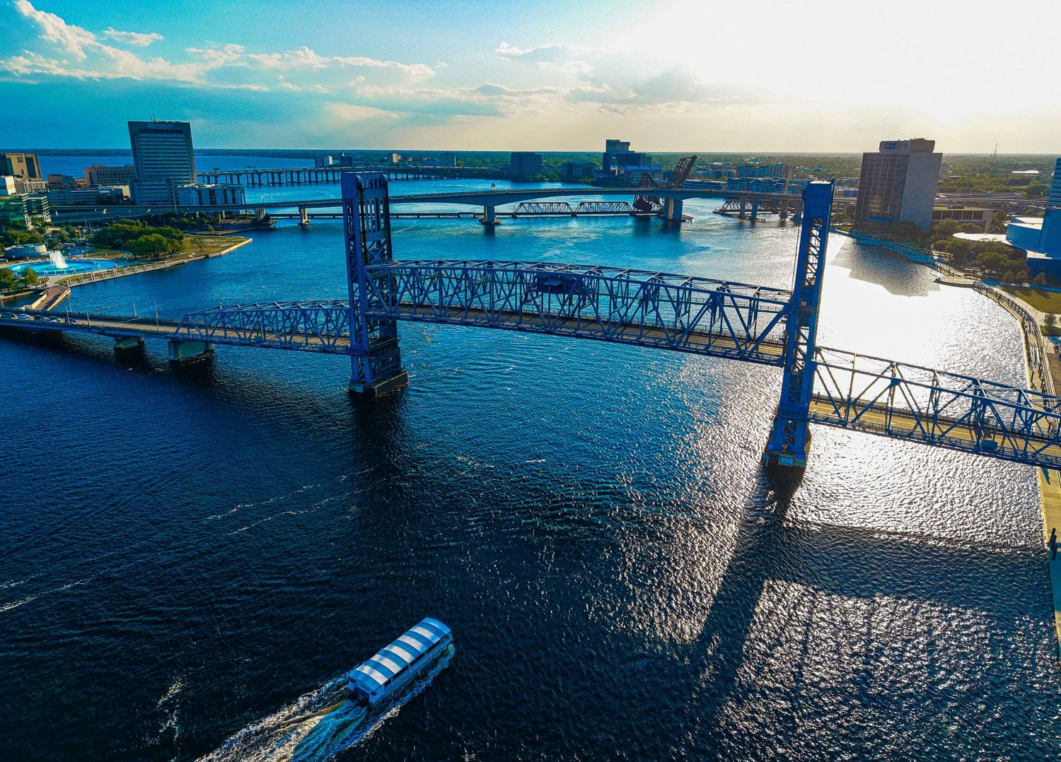Aerial view of a blue steel lift bridge over a sunlit river with a boat passing beneath, city skyline in background.