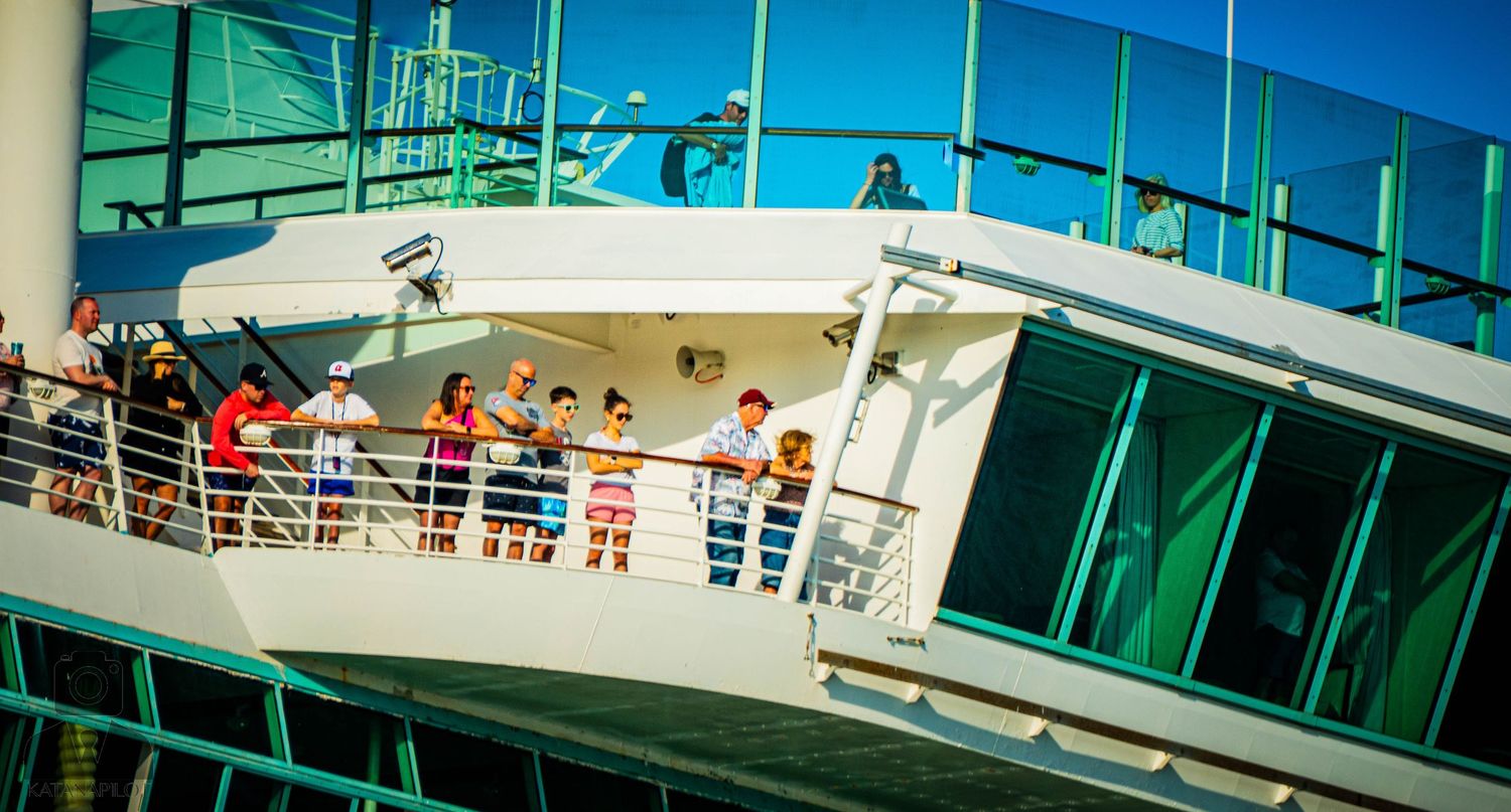Passengers gather on the upper deck of a cruise ship with turquoise glass panels on a sunny day at sea.
