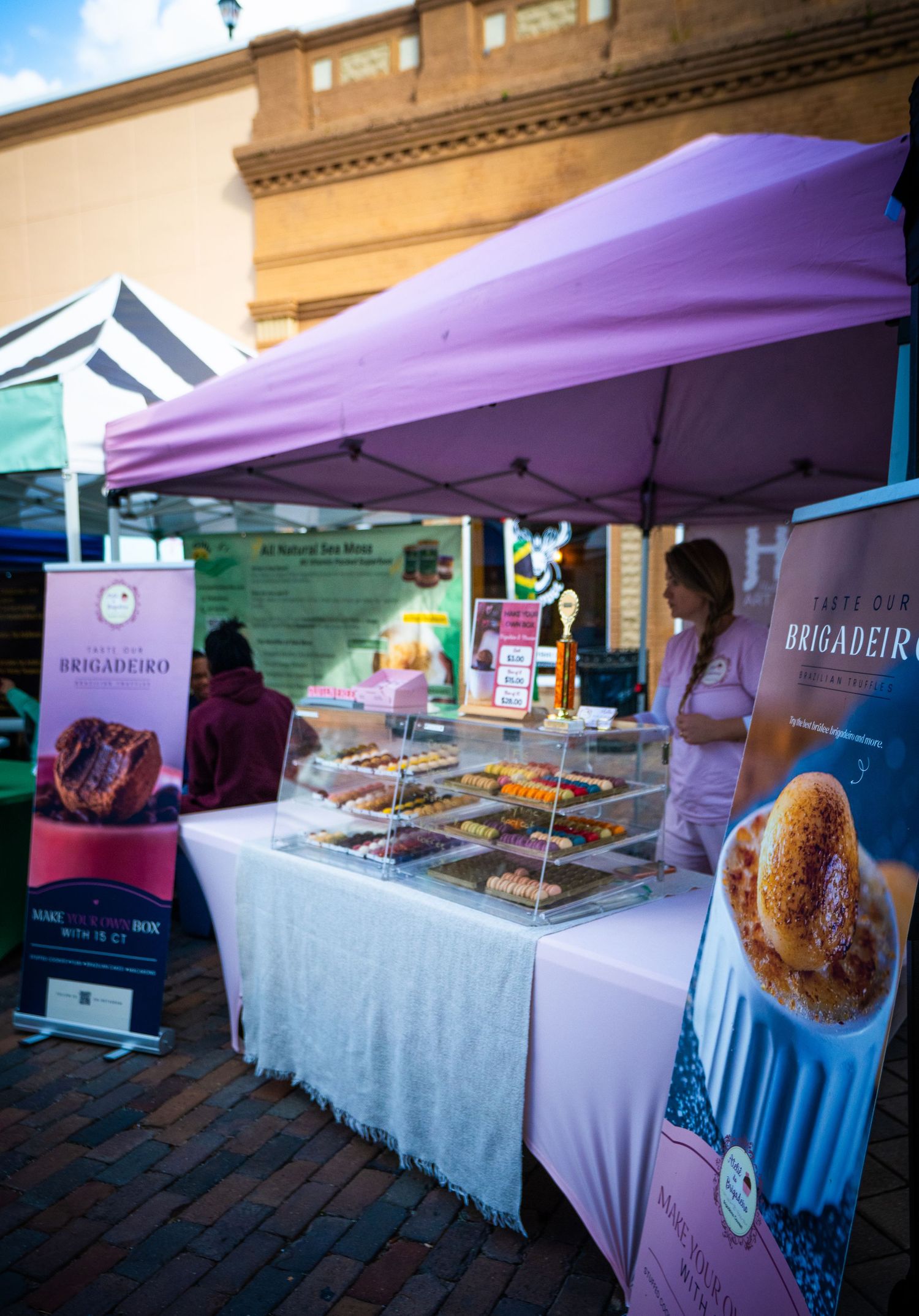 Farmers market vendor booth with purple canopy displays fresh baked goods and treats on white-clothed tables.
