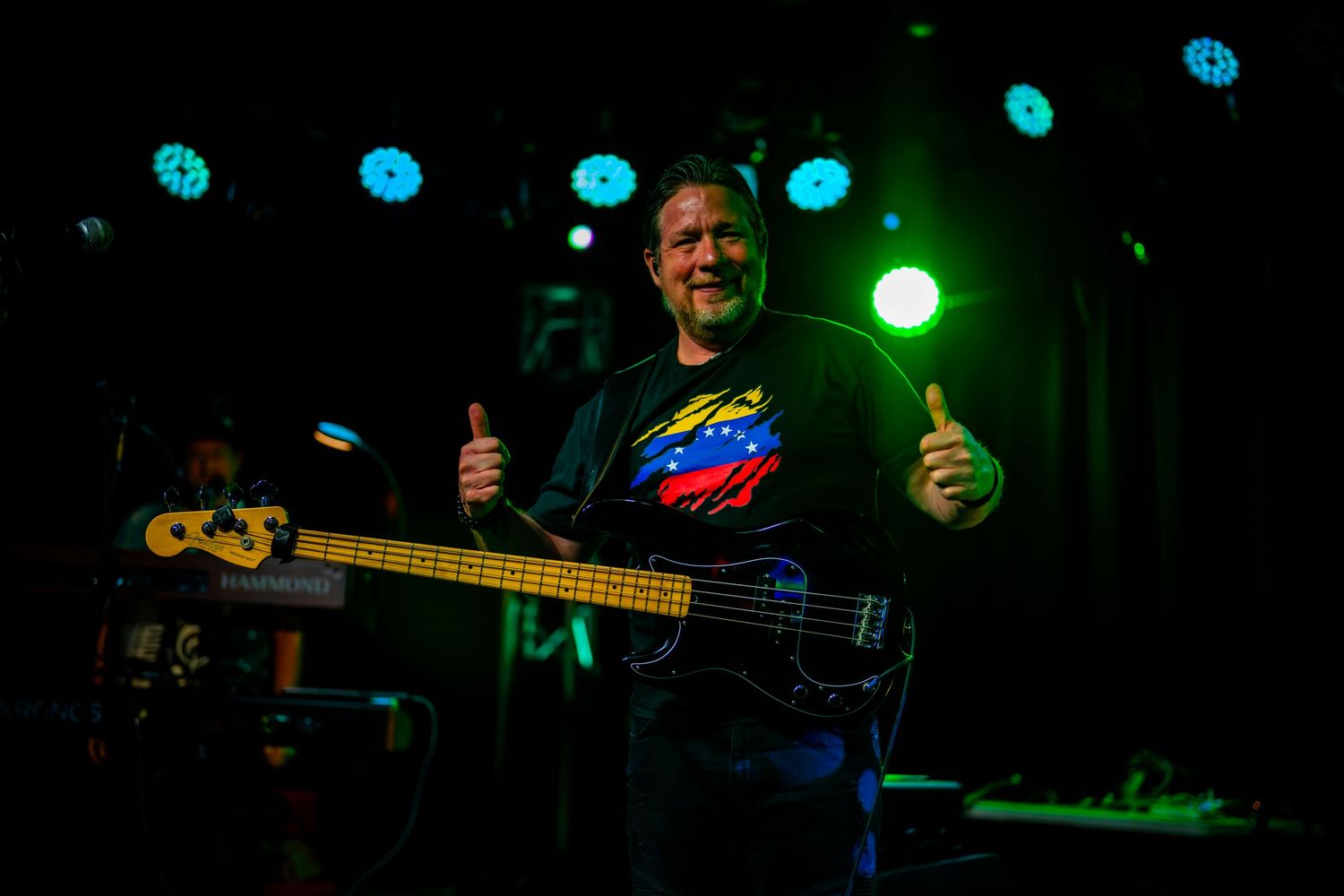 Smiling musician gives thumbs up on stage holding bass guitar under colorful concert lights.