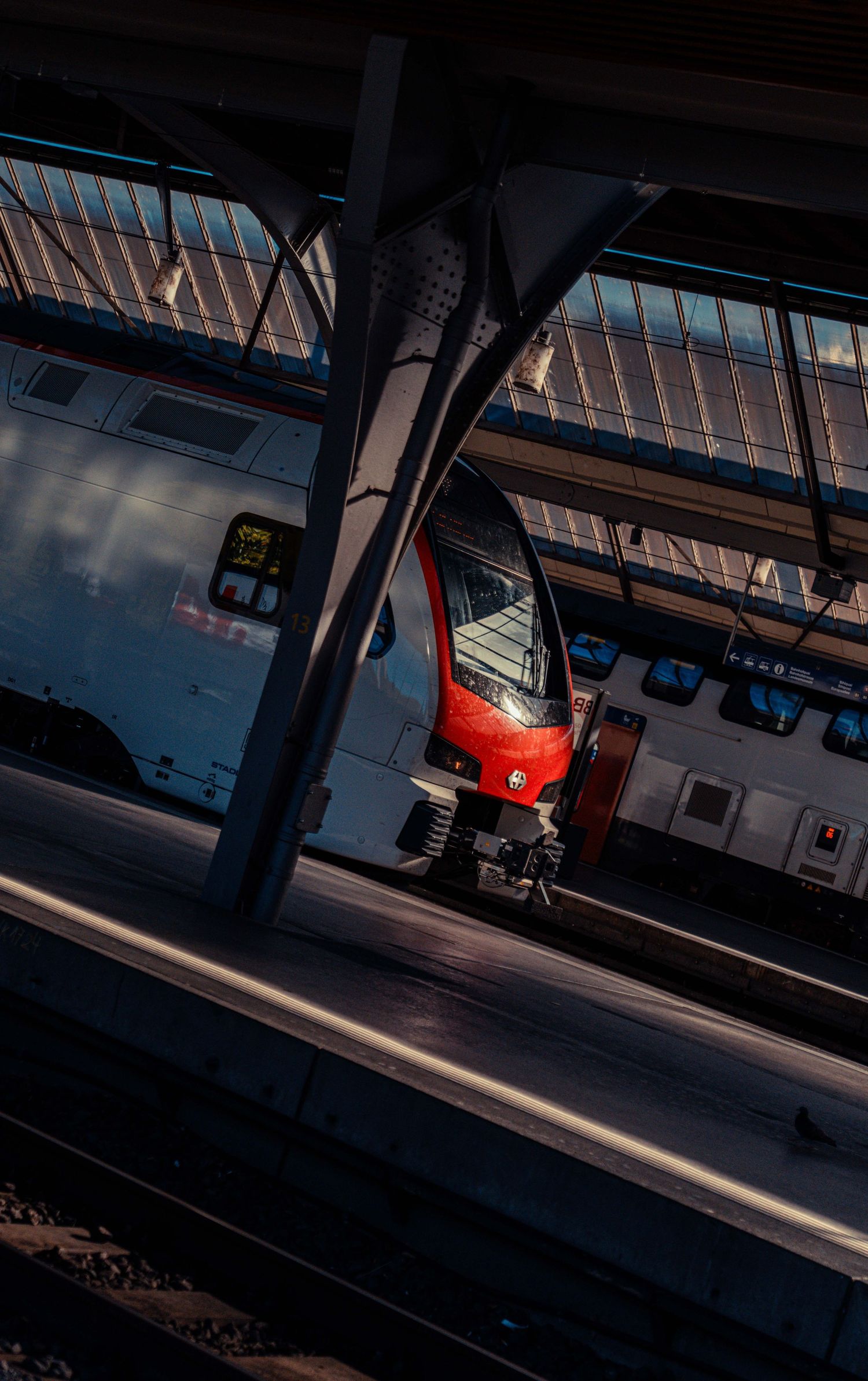 Red and white passenger train waiting at a platform beneath a steel-framed glass roof in a train station, lit by warm evening sunlight.