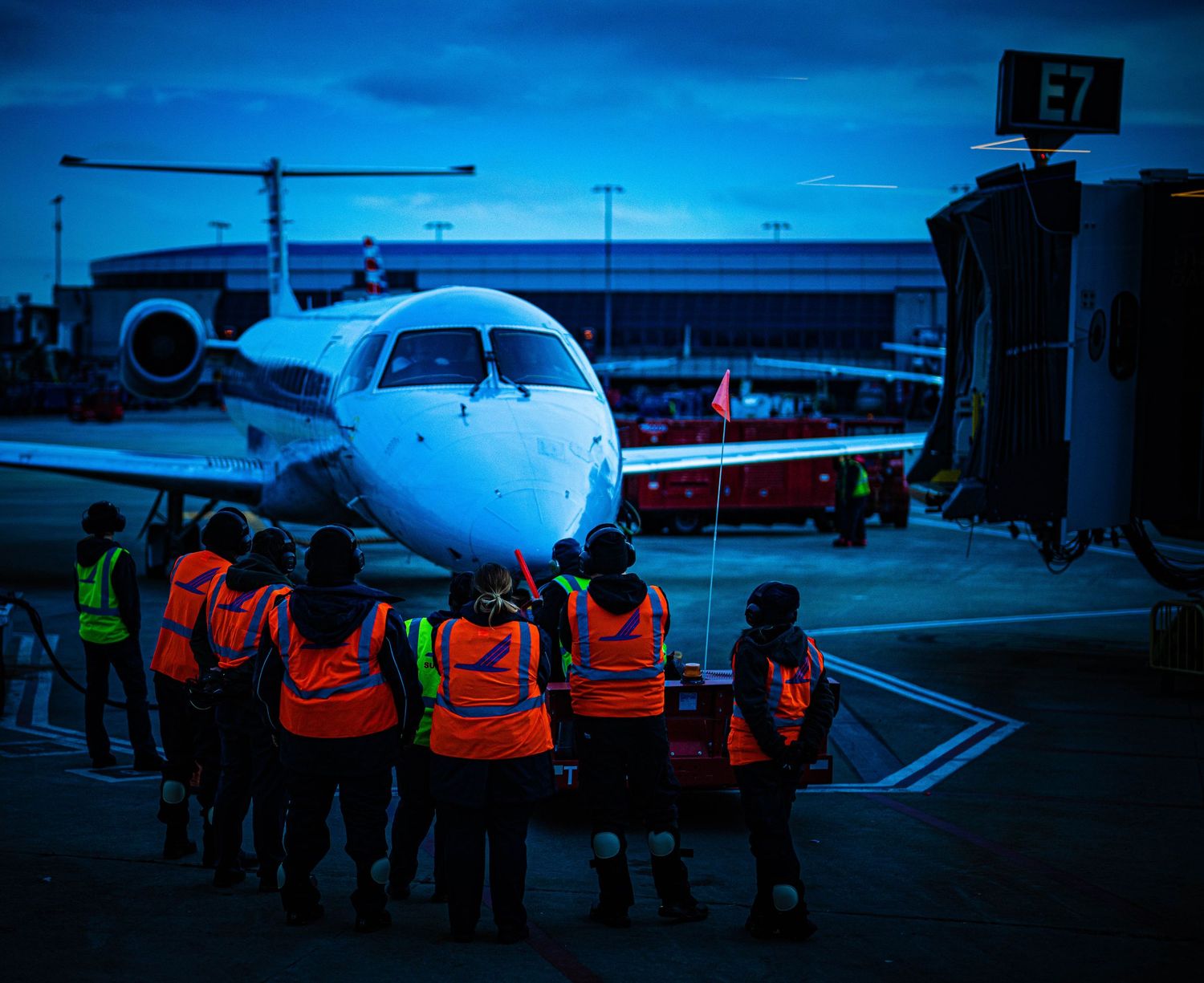 Ground crew in orange safety vests prepare an aircraft for departure on a dark tarmac at dusk.