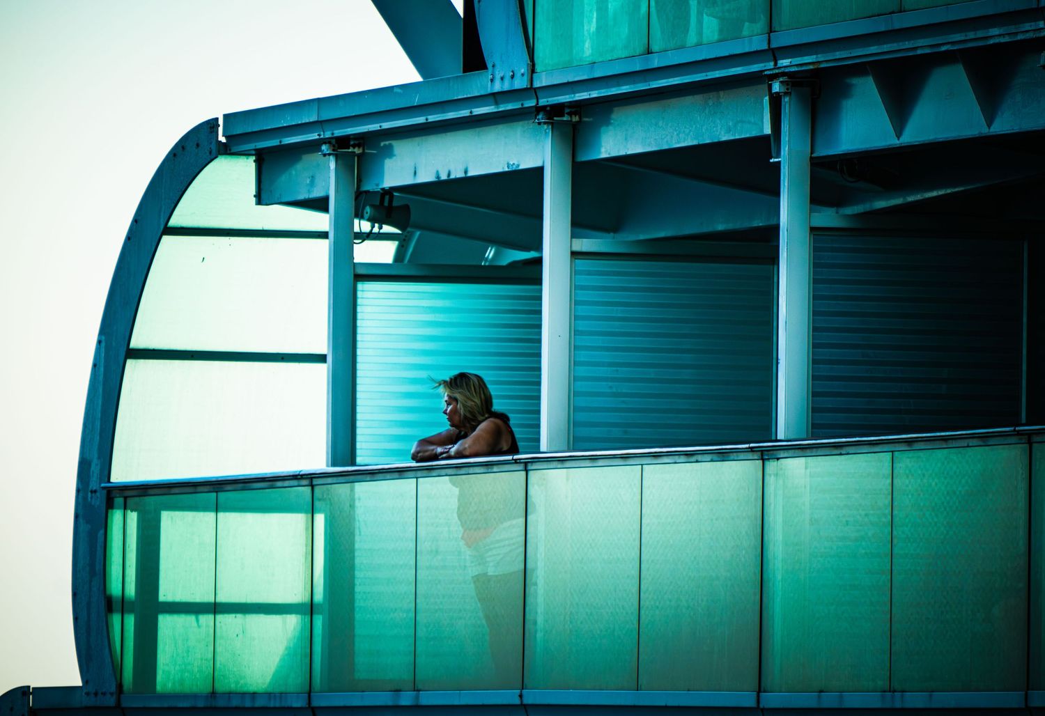 A person sits in a curved glass window alcove with turquoise-tinted lighting creating a moody architectural atmosphere.