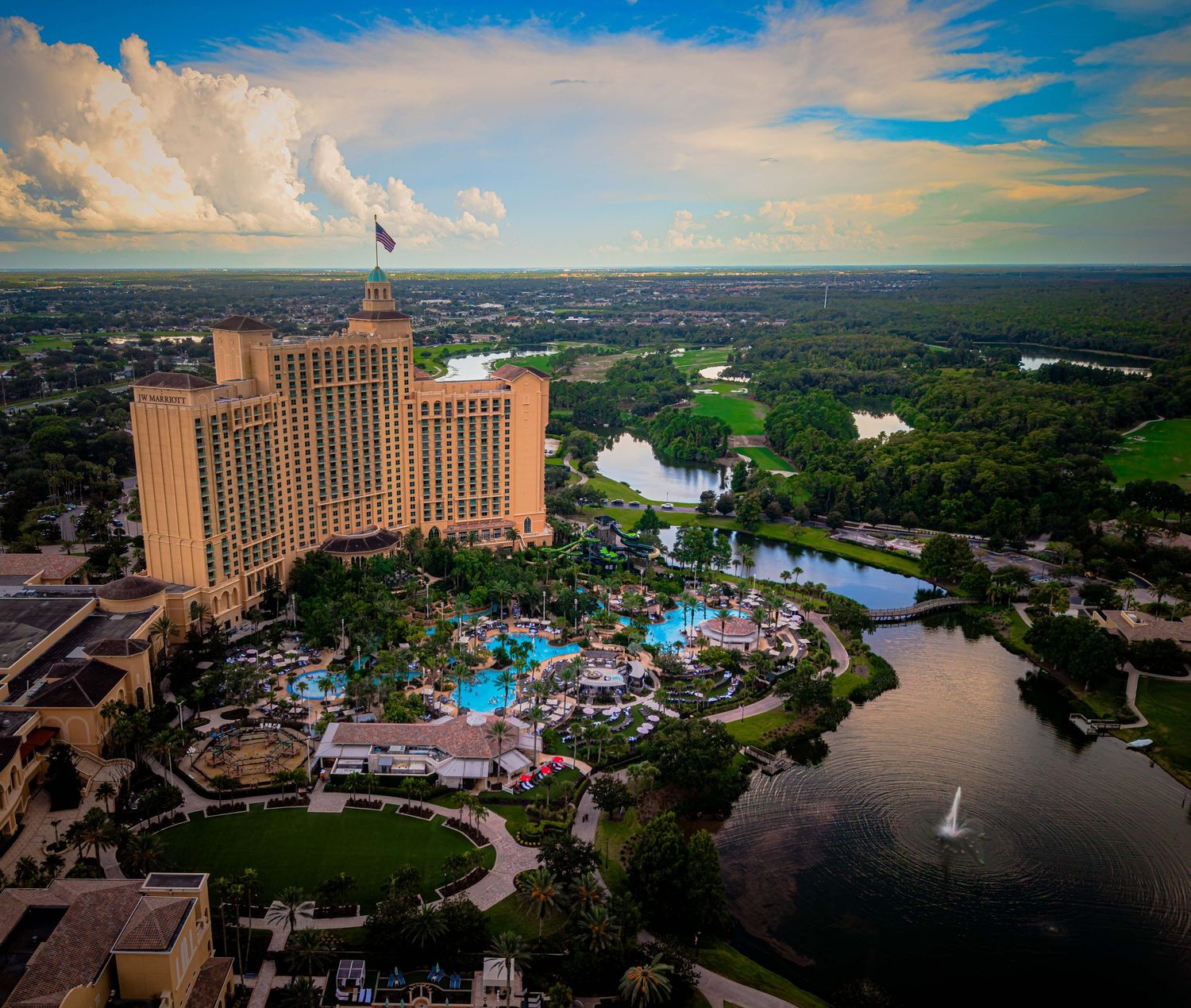 Aerial view of a luxury resort hotel with pools, lush gardens, and a scenic lake surrounded by greenery under a dramatic sky.