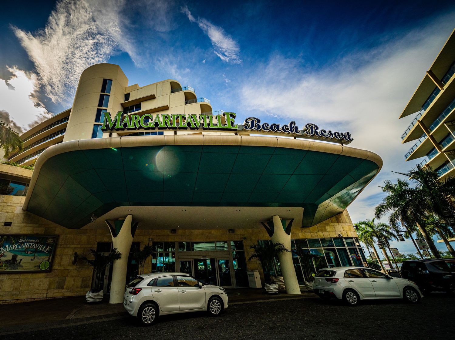 Margaritaville Beach Resort exterior entrance with palm trees, luxury cars, and vibrant signage under a blue sky.