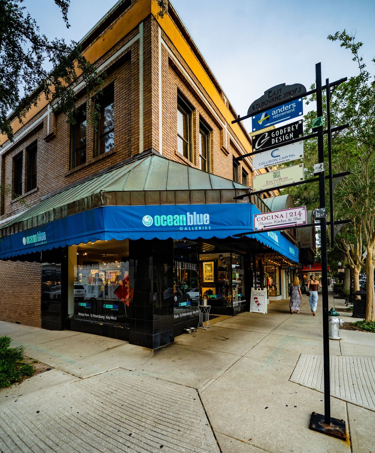 Exterior view of a shopping plaza with blue awnings and brick sidewalks.