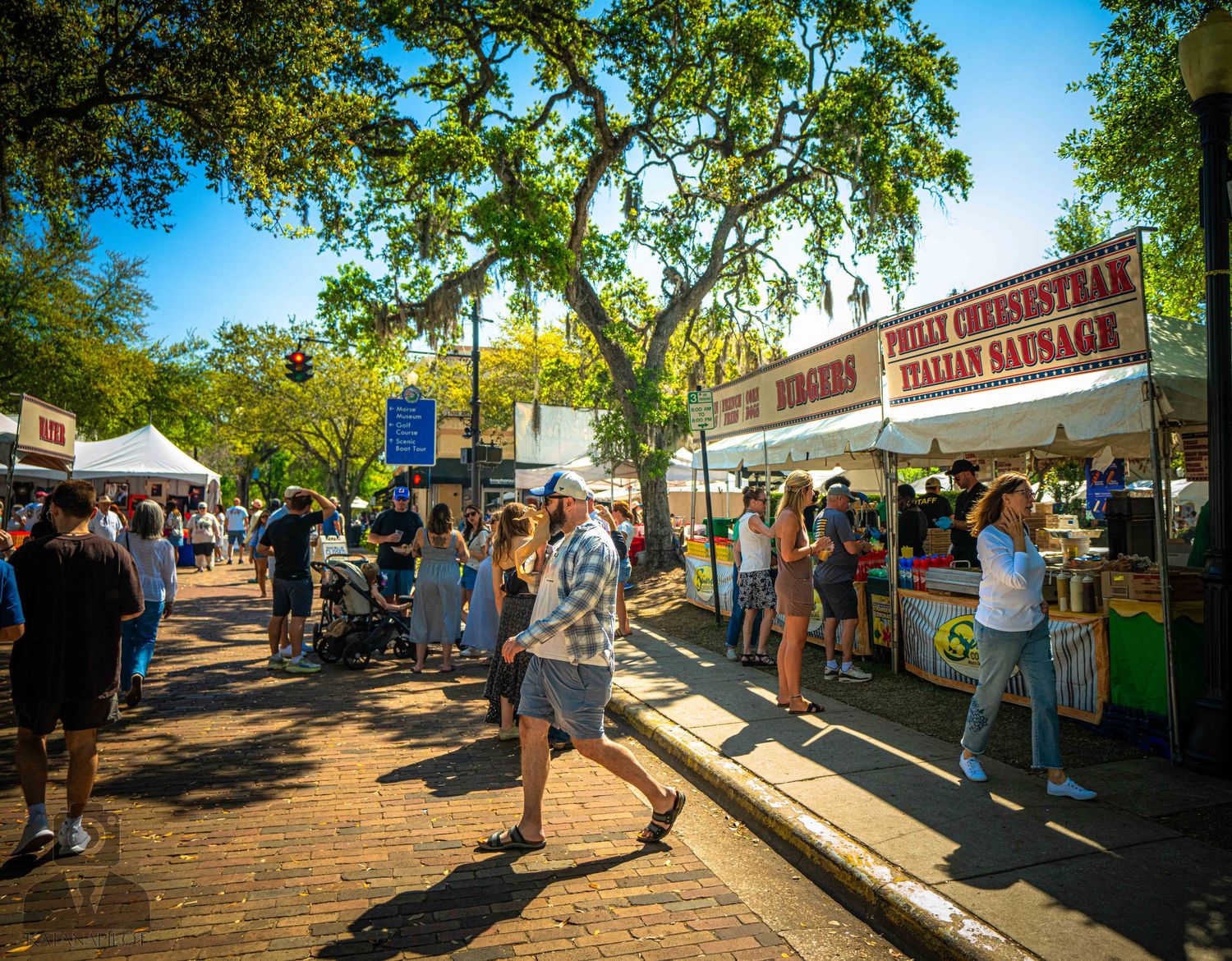 Visitors browse food vendor stalls at an outdoor market under shady trees on a sunny afternoon.