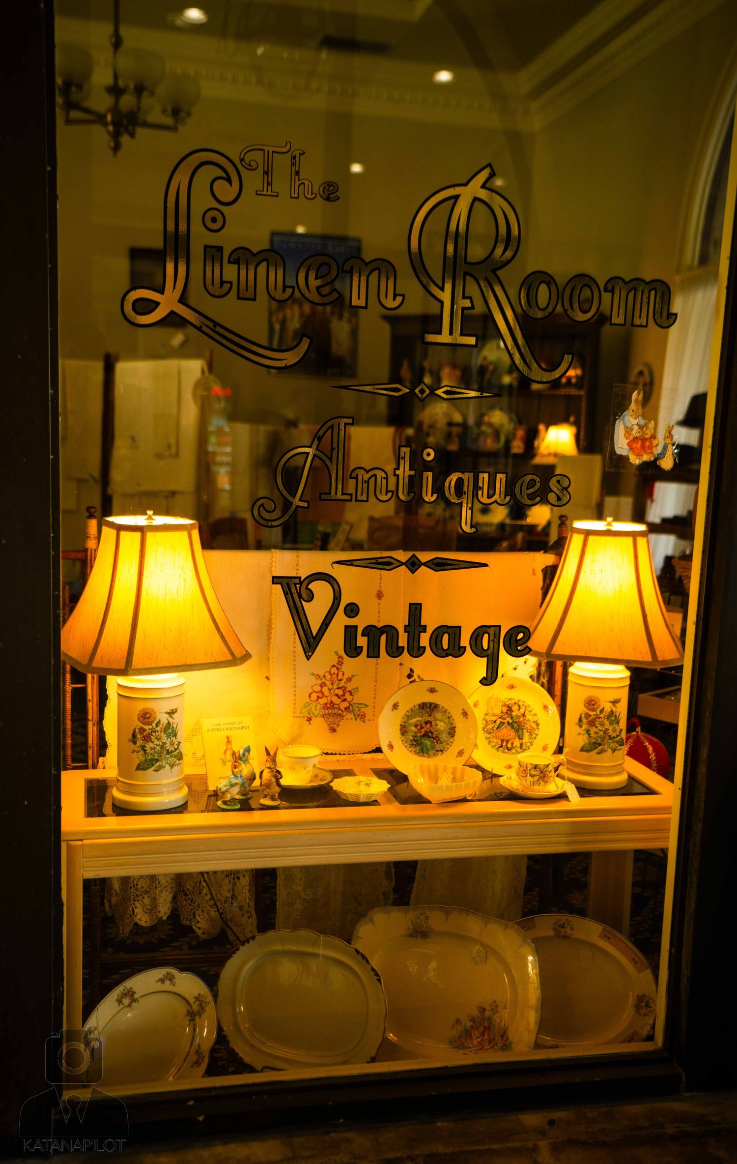 Illuminated vintage display window showcases antique china and glassware under warm lamp lighting at night.