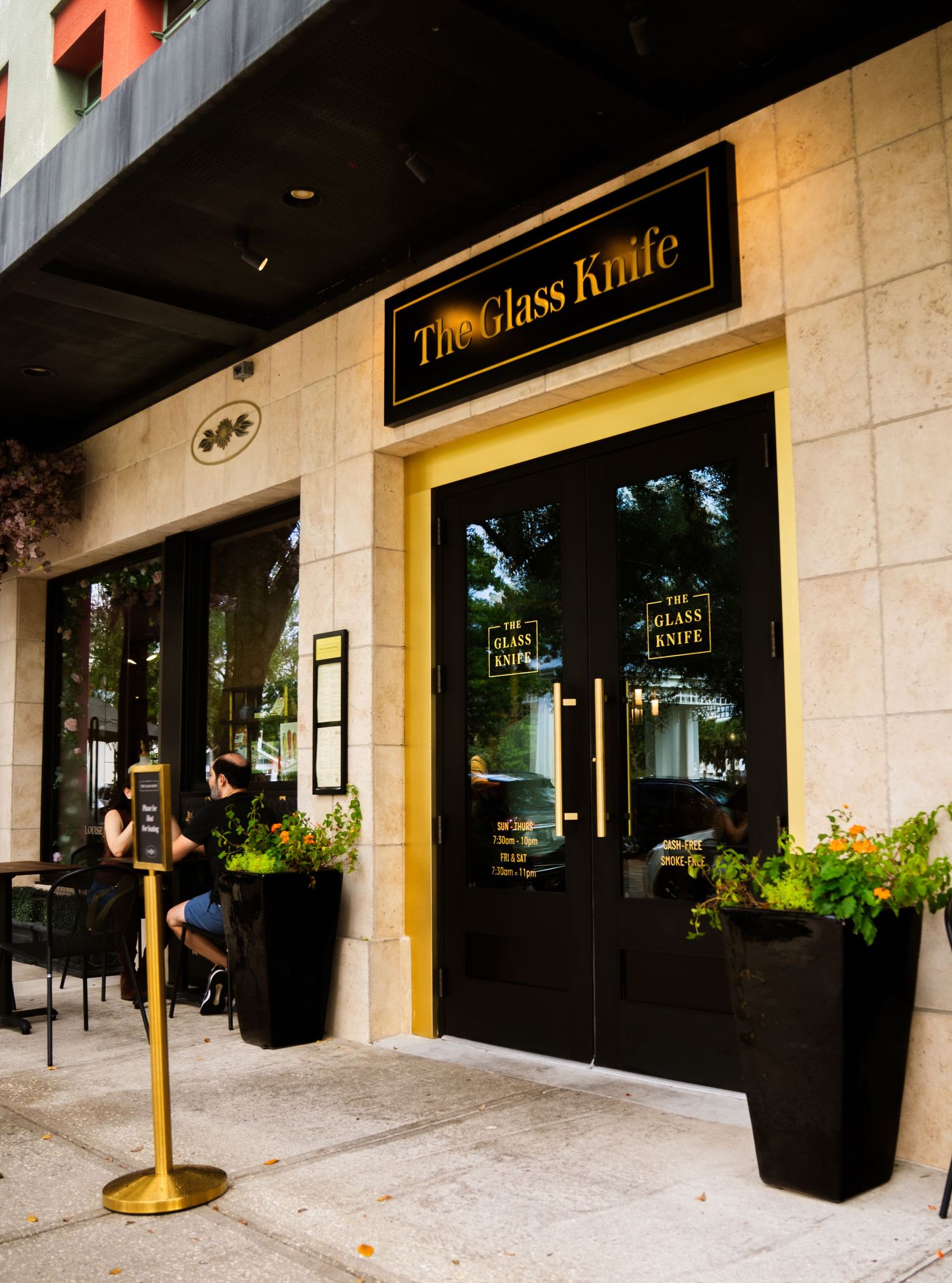 Exterior view of an upscale restaurant or cafe storefront with planters and gold trim details.