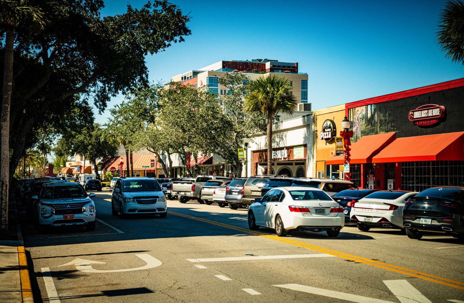 A panoramic view of a suburban shopping center parking lot with cars and storefronts under a bright blue sky.
