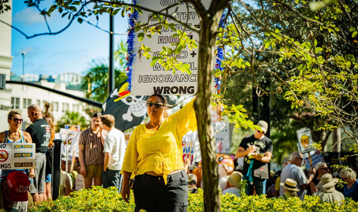 Protesters gather on a sunny day holding colorful signs during a street demonstration.