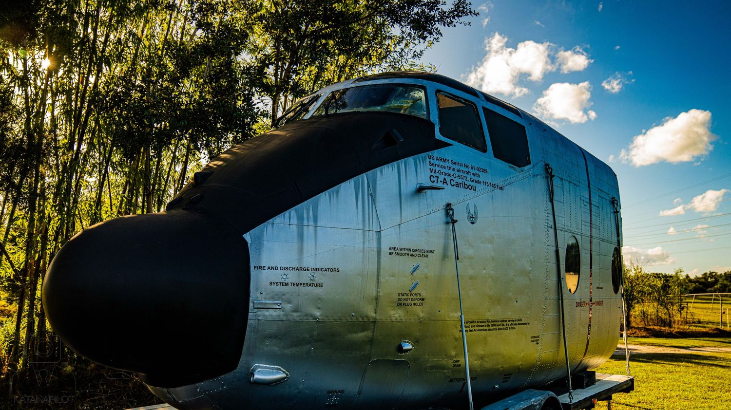 Silver vintage aircraft on display in outdoor setting with trees and clouds in background.