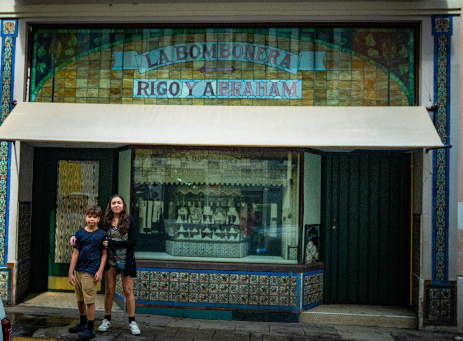 Two people stand in front of a vintage storefront with decorative tiles and a sign reading 'Rico y Abeijan'.