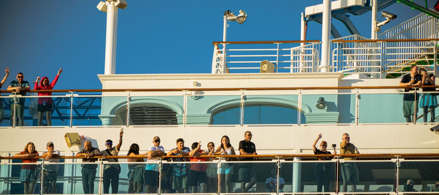 Passengers gather along the railing of a cruise ship deck against a bright blue sky on a sunny day at sea.