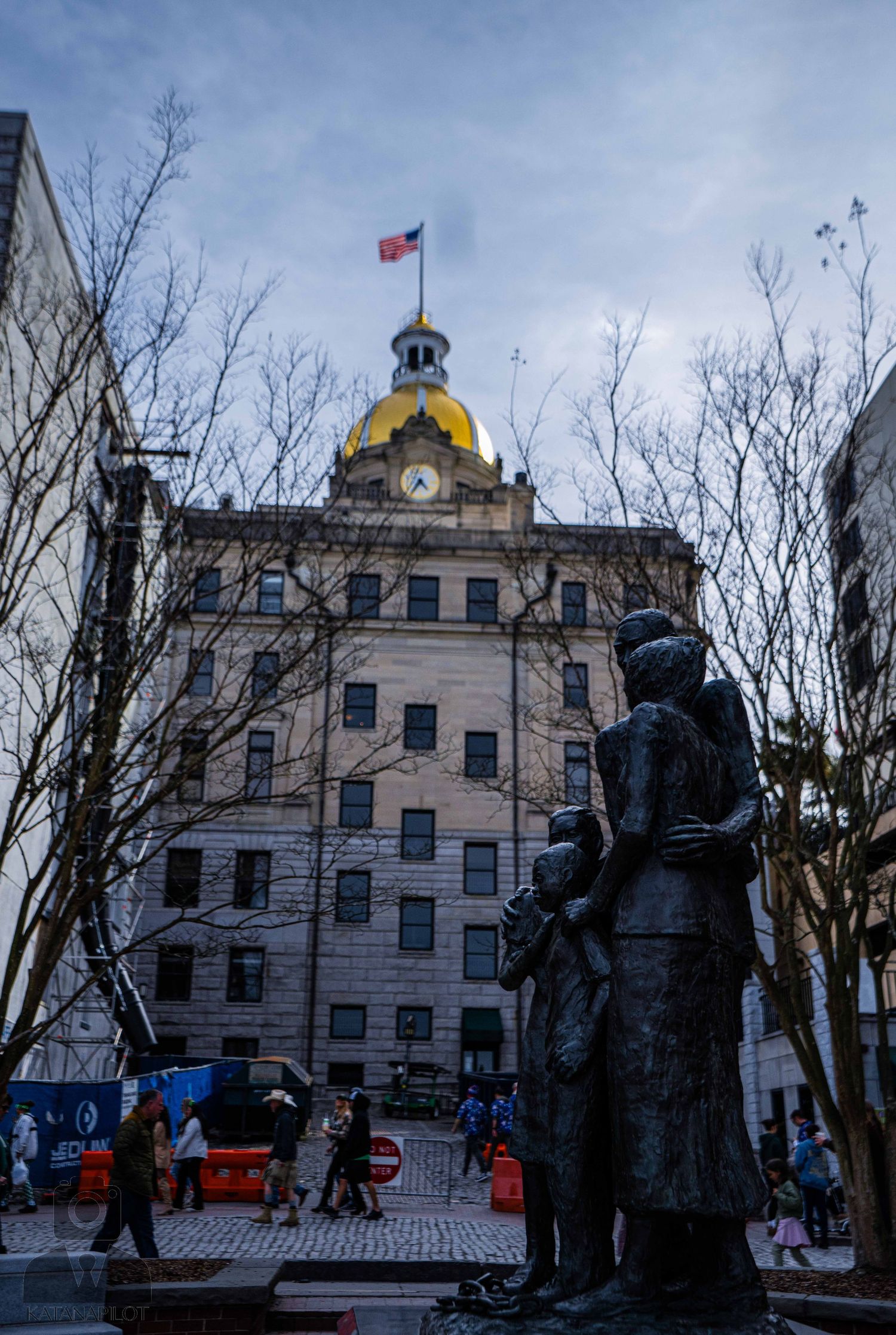 Row of bronze statues stand in front of a grand government building with a golden dome on a winter day.