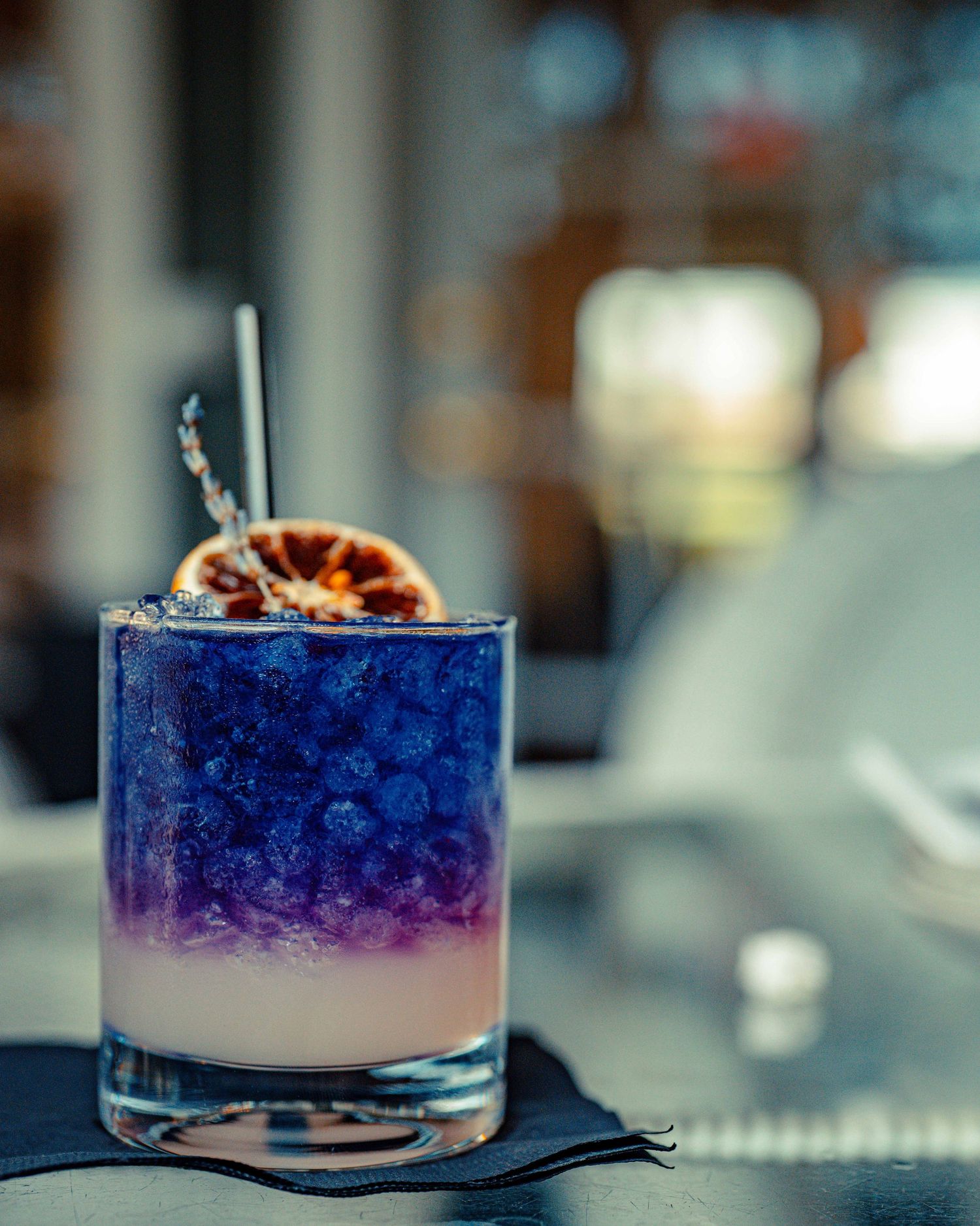 A layered cocktail with blue and purple gradient colors garnished with a dried citrus wheel and straw in a rocks glass.