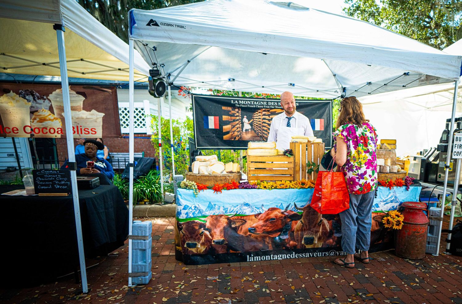 Vendor booth at outdoor market displays colorful products under white tent canopy on brick pavement.