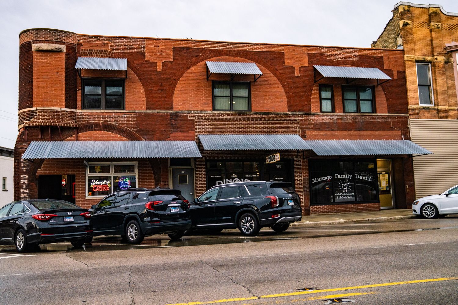 Continuous streetscape of connected brick commercial buildings with parked cars in a downtown setting.