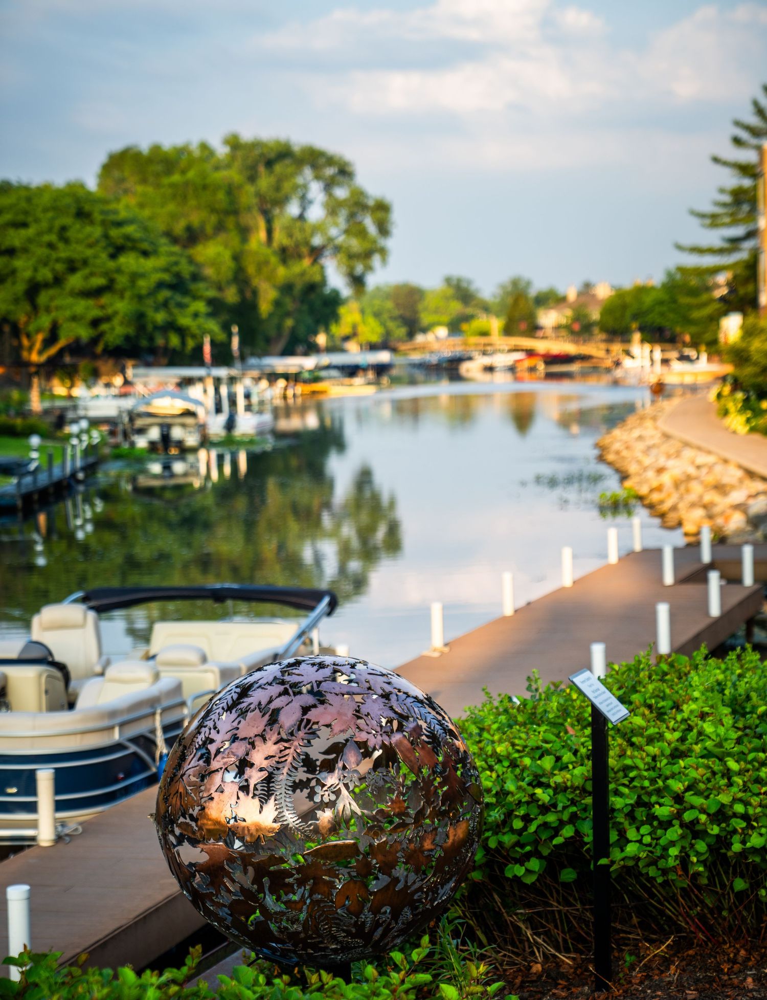 Curved brick walkway along a tranquil waterfront with decorative metal spheres and white posts at sunset.