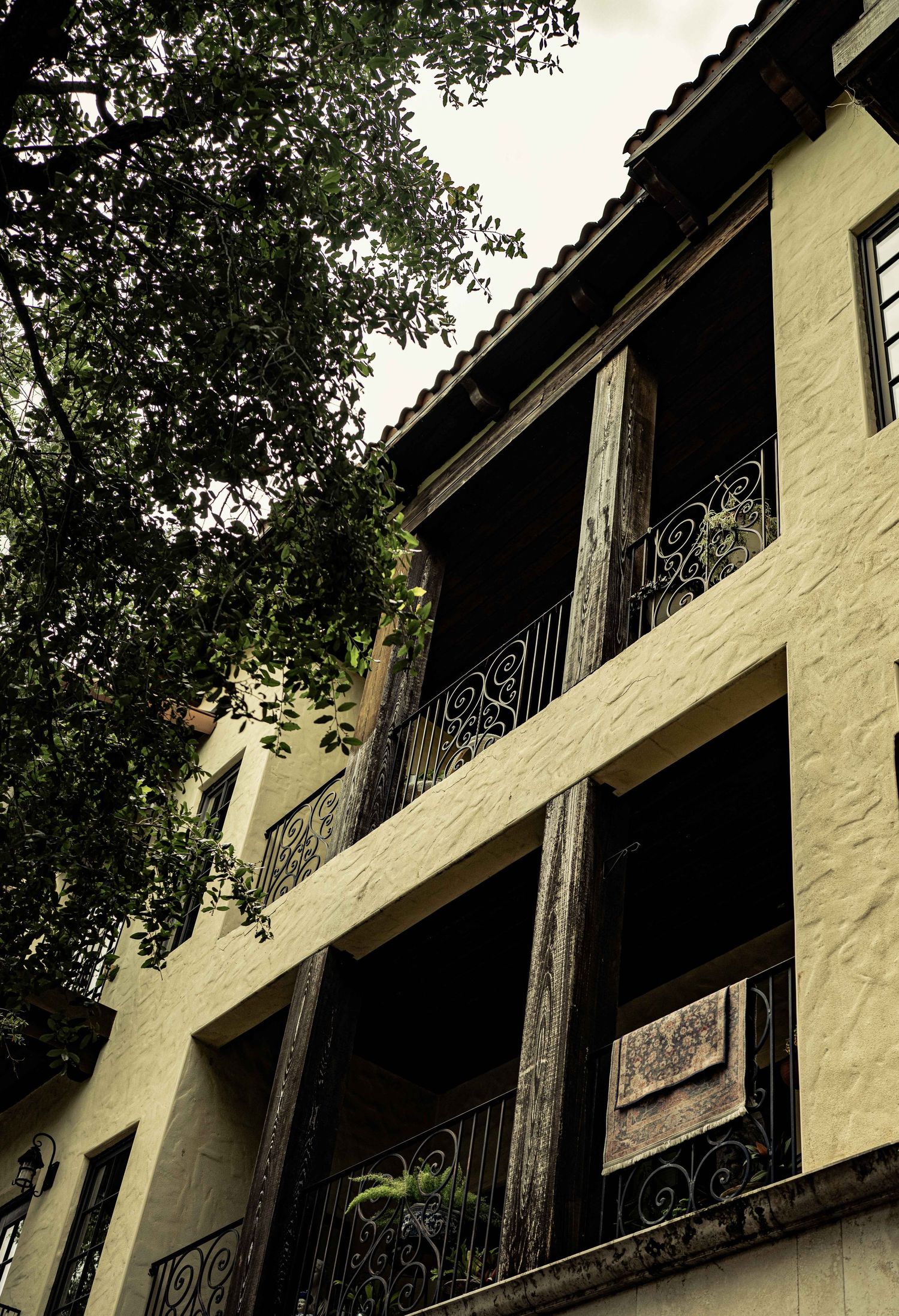 Historic building with ornate wrought iron balconies, wooden columns, and yellow stucco walls surrounded by lush trees.