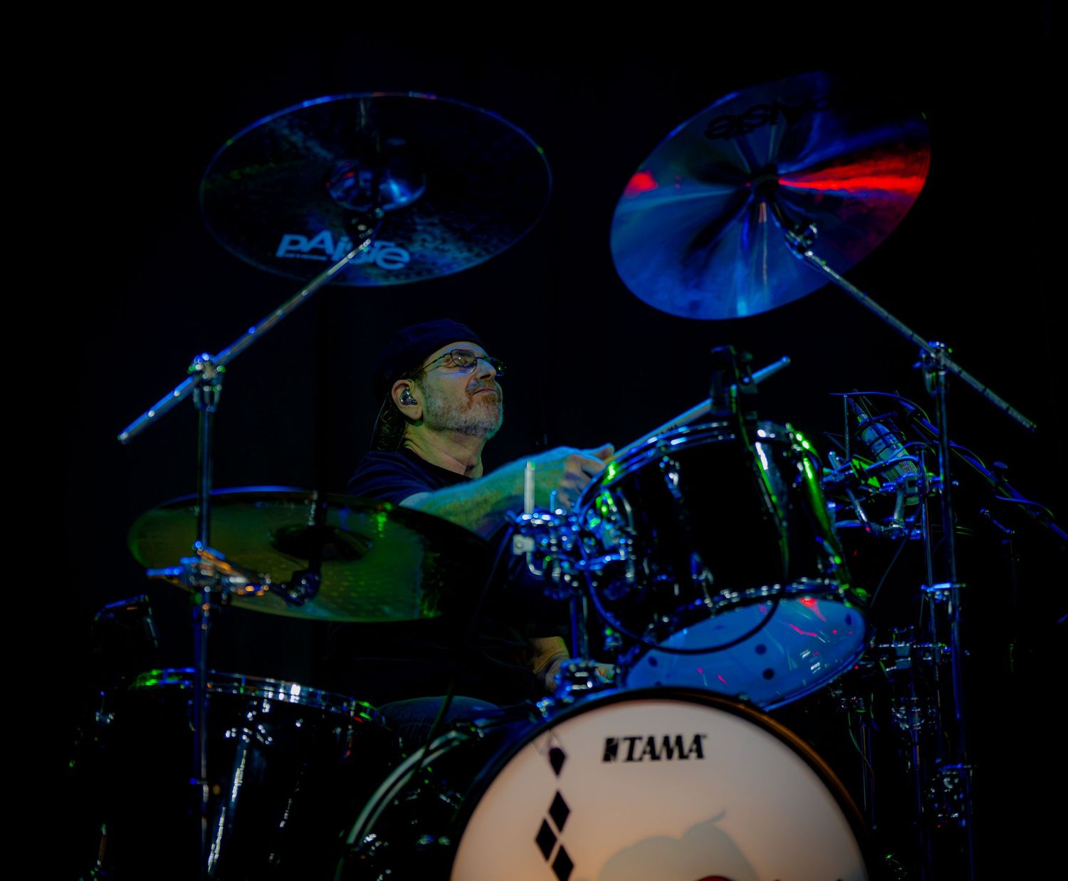 Musician playing Tama drum kit on stage with cymbals illuminated by blue and green stage lights.