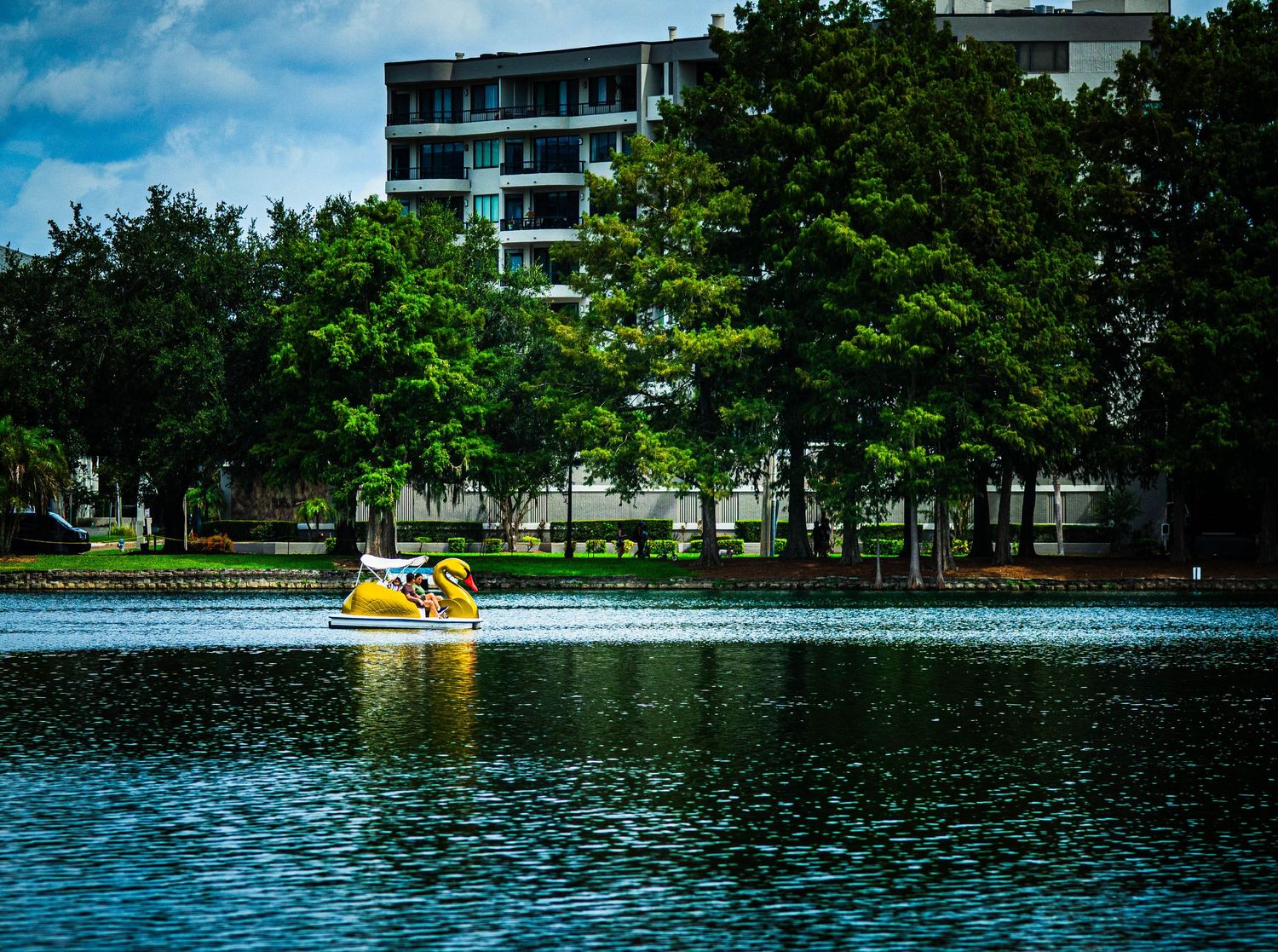Yellow paddle boat on calm lake with lush green trees and modern apartment building in background.