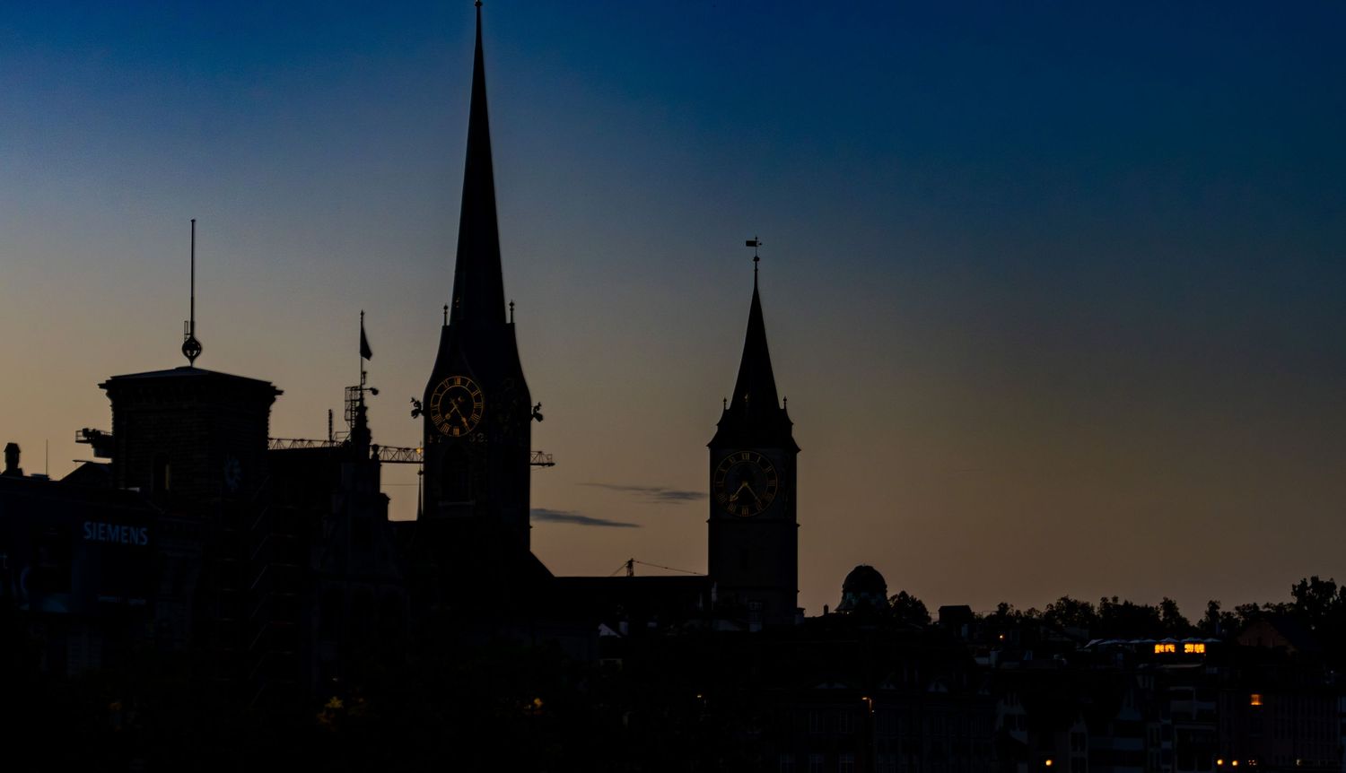 Silhouette of church spires and a clock tower against a deep blue and orange twilight sky over a darkened city skyline at dusk.
